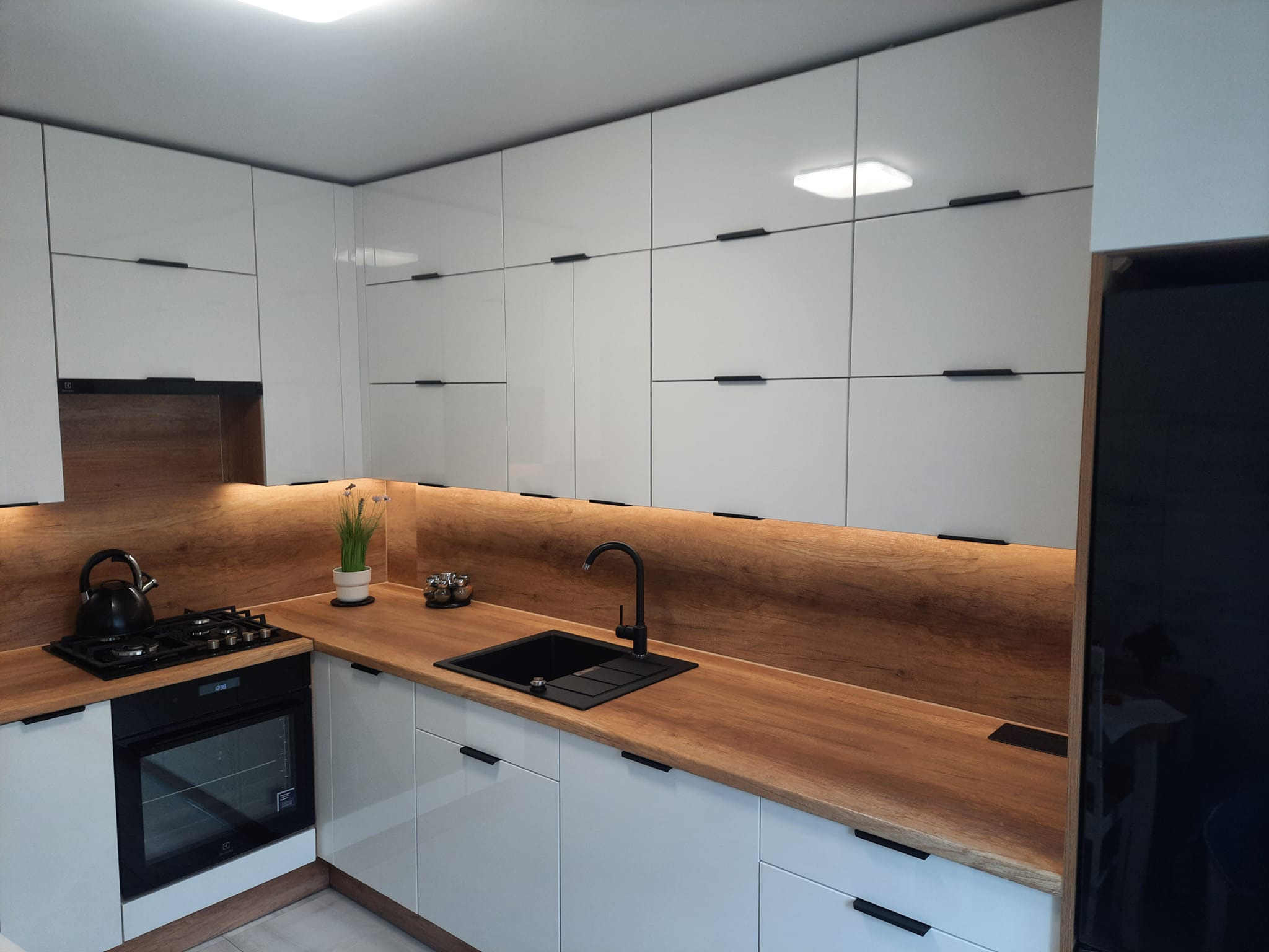 Nowoczesna kuchnia z białymi, lakierowanymi szafkami, drewnianym blatem i czarnymi akcentami: bateria, zlewozmywak, uchwyty, płyta grzewcza i czajnik.