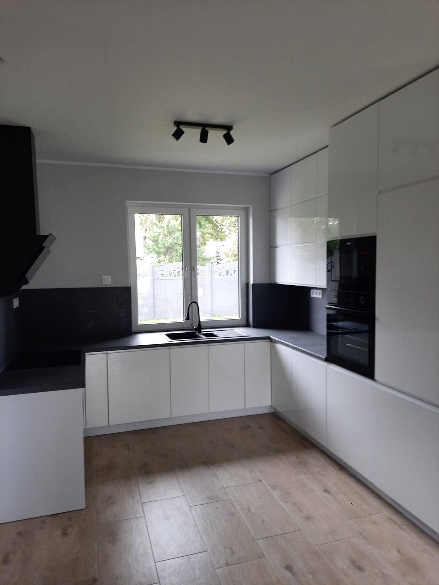 Nowoczesna kuchnia z białymi, lakierowanymi frontami szafek, czarnym blatem i sprzętem AGD, widok na okno z zielenią na zewnątrz, drewnopodobna podłoga z płytek.