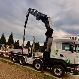 HDS Niepolomice - Ciężarówka z dźwigiem HIAB i platformą transportową przewozi rury oraz koparkę na tle pochmurnego nieba i zieleni. Widoczny element dekoracyjny na kabinie.