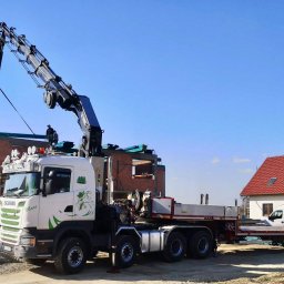 HDS Niepolomice - Ciężarówka z dźwigiem podnosi element konstrukcyjny na budowie domu jednorodzinnego. W tle widoczny ukończony budynek z czerwoną dachówką.