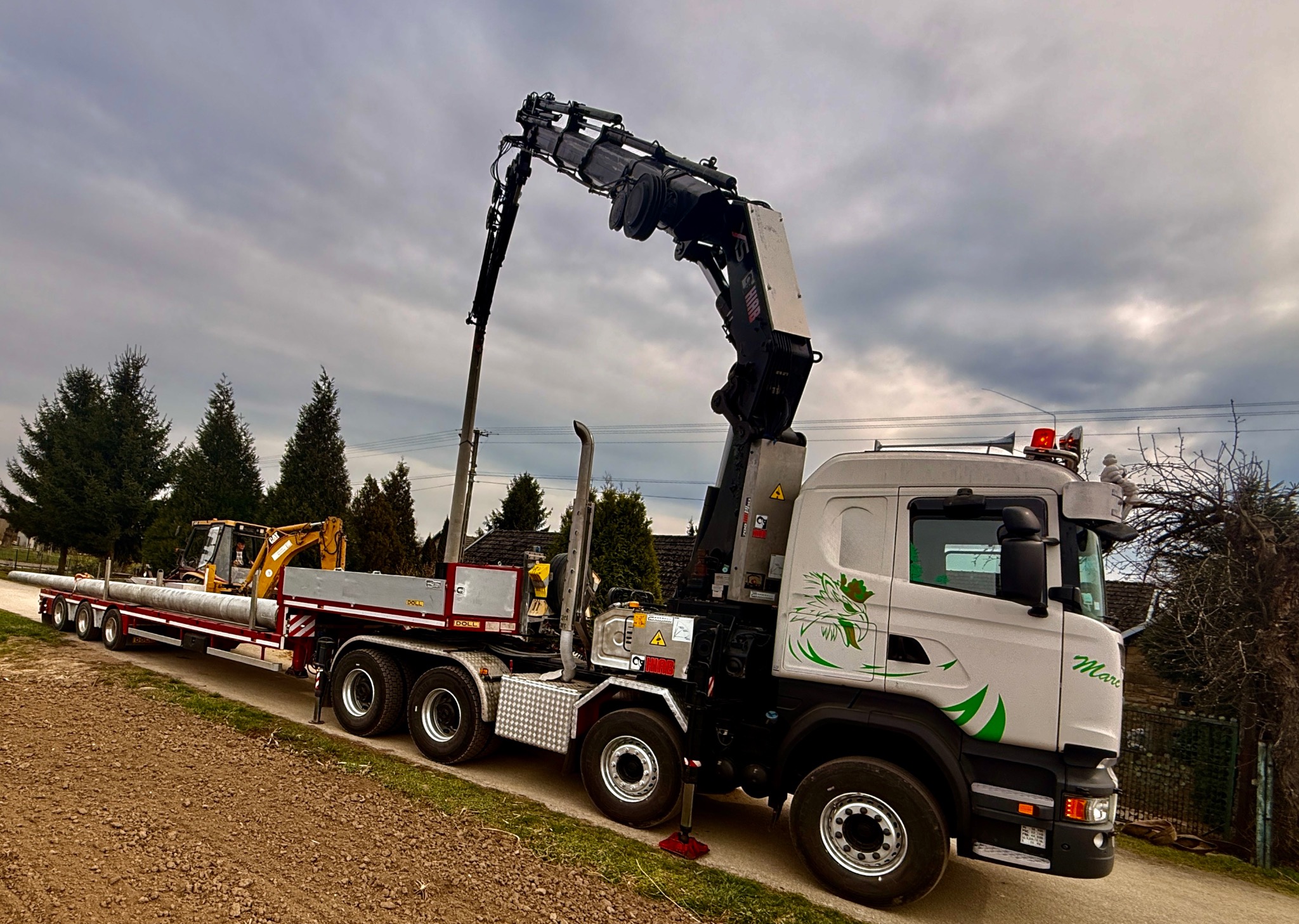 Ciężarówka z dźwigiem HIAB i platformą transportową przewozi rury oraz koparkę na tle pochmurnego nieba i zieleni. Widoczny element dekoracyjny na kabinie.