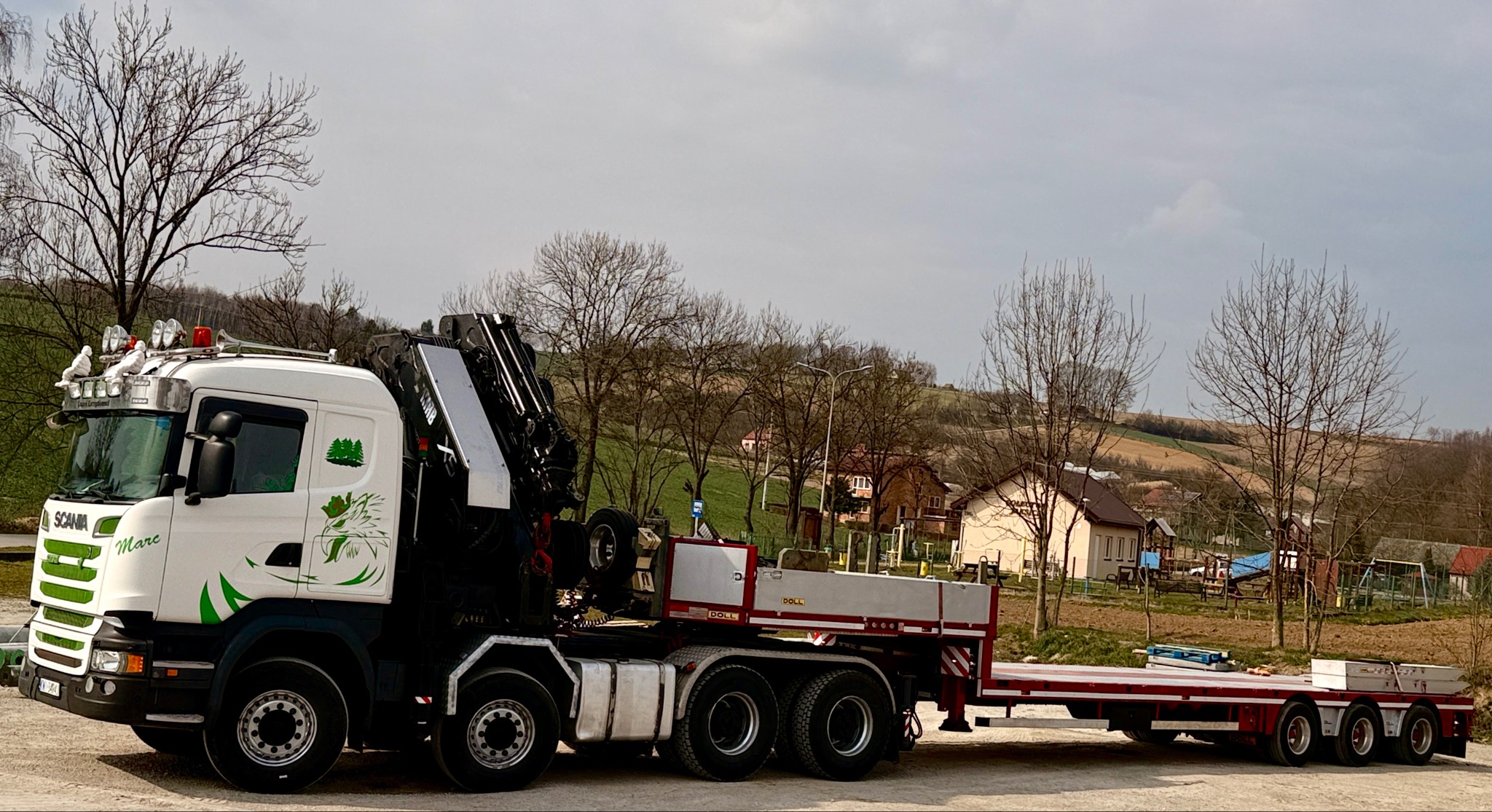 Biała laweta Scania z żurawiem i przyczepą na tle wiejskiego krajobrazu. Na kabinie ozdobne figurki gołębi i zielone grafiki. Widoczne detale konstrukcji i oświetlenia.
