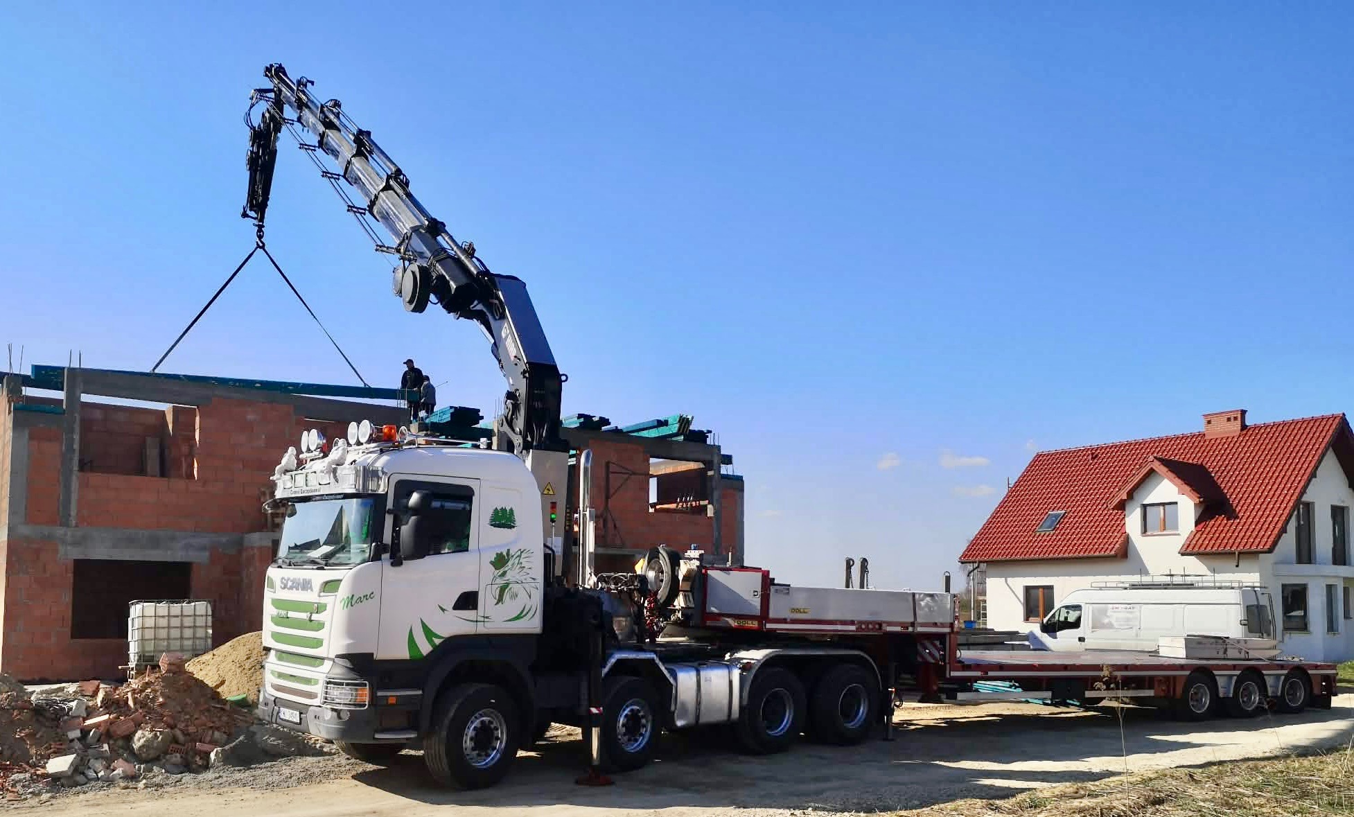 Ciężarówka z dźwigiem podnosi element konstrukcyjny na budowie domu jednorodzinnego. W tle widoczny ukończony budynek z czerwoną dachówką.
