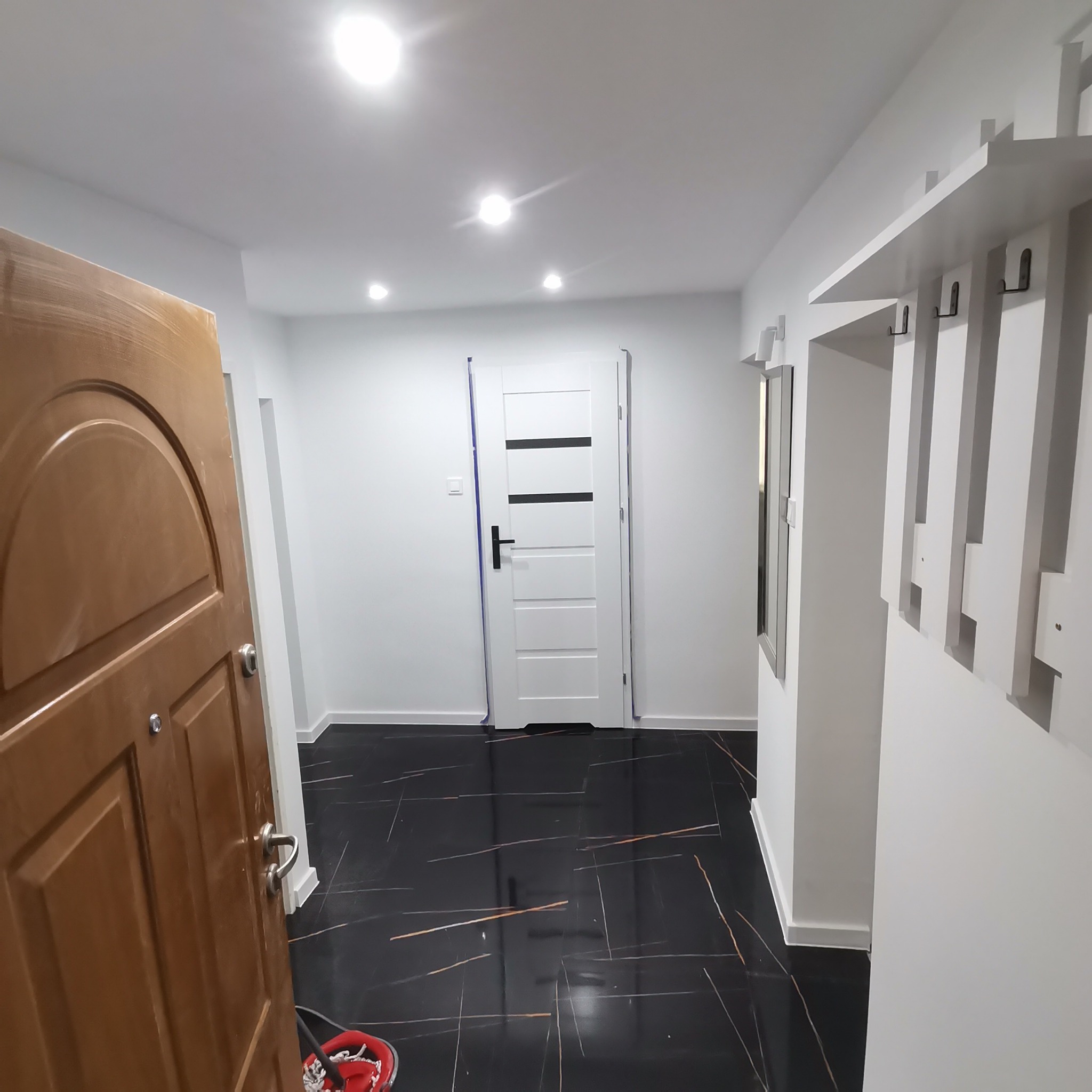 Nowoczesny przedpokój z czarną podłogą imitującą marmur, białe ściany, drewniane drzwi wejściowe, białe drzwi wewnętrzne z czarnymi paskami i minimalistyczny wieszak z lustrem.