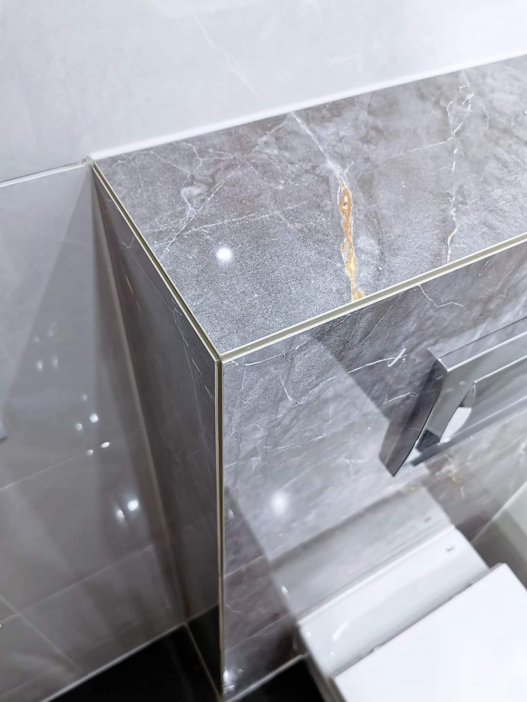 Szara marmurowa półka w łazience z delikatnym złotym żyłkowaniem, wykończona cienką złotą listwą, zamontowana nad toaletą, z widocznym fragmentem chromowanego uchwytu na ręczniki.