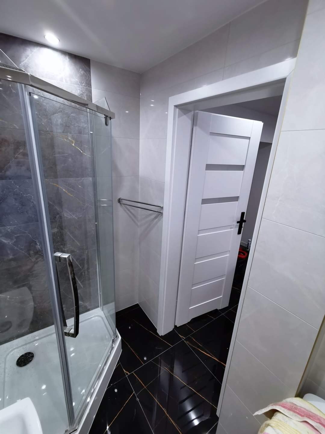 Nowoczesna łazienka z kabiną prysznicową ze szkła hartowanego, czarną podłogą z żyłkowaniem i białymi płytkami ściennymi, widok na białe drzwi z poziomymi panelami.