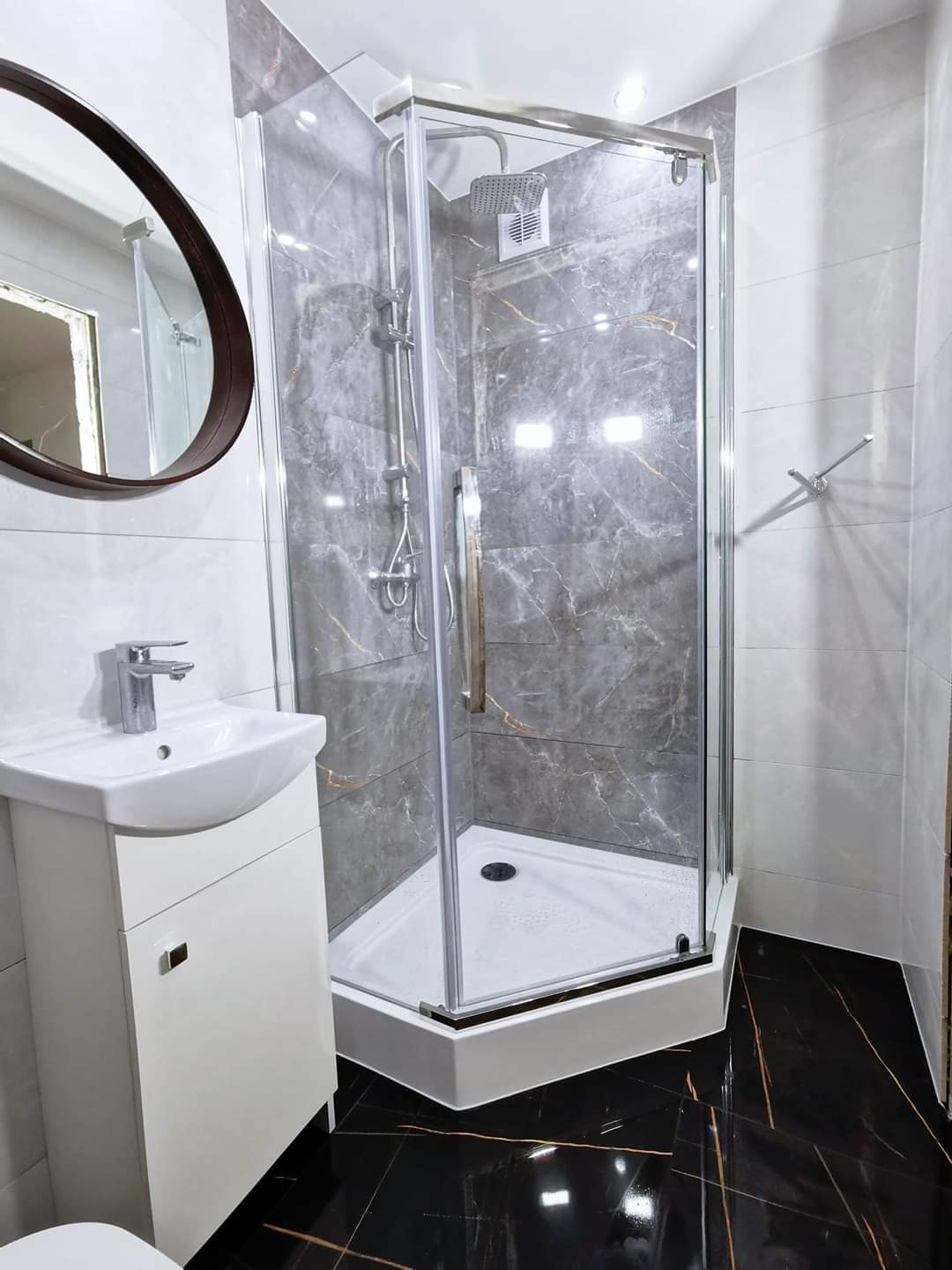 Nowoczesna łazienka z kabiną prysznicową wyłożoną marmurkowymi płytkami, białą umywalką z szafką i czarną, błyszczącą podłogą z żyłami. Okrągłe lustro z ciemną ramą.