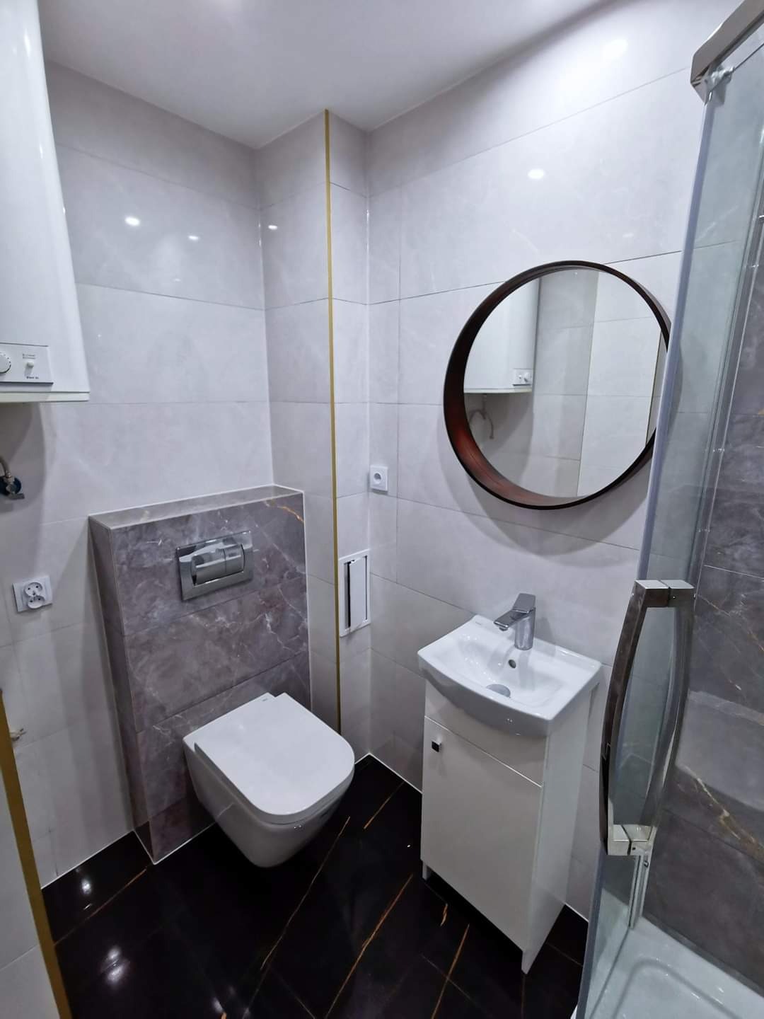 Nowoczesna łazienka po remoncie: biała umywalka z szafką, wisząca toaleta obudowana marmurową płytą, okrągłe lustro w ciemnej ramie, czarne płytki podłogowe z złotymi żyłkami, kabina prysznicowa...