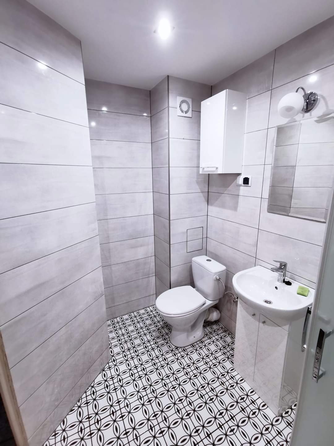 Nowoczesna łazienka po remoncie: szare płytki imitujące drewno na ścianach, wzorzysta czarno-biała podłoga, biała umywalka z szafką, toaleta i szafka wisząca.