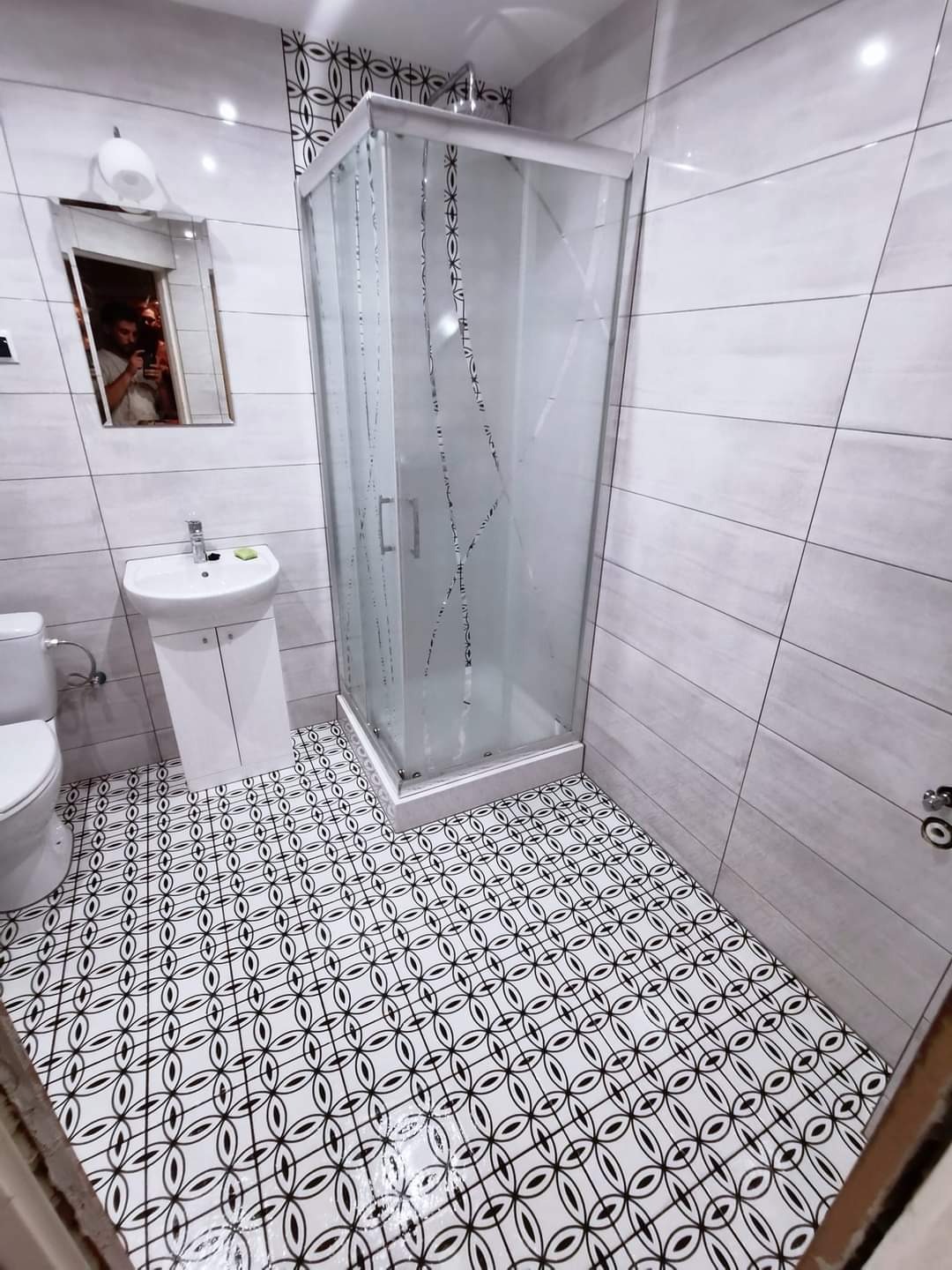 Nowoczesna łazienka z kabiną prysznicową, białą umywalką na szafce i wzorzystą czarno-białą podłogą. W lustrze odbicie osoby robiącej zdjęcie telefonem.