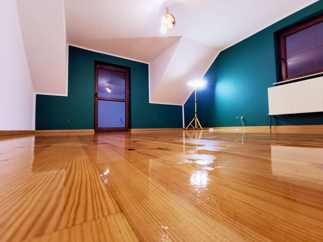 Błyszcząca, lakierowana podłoga z jasnego drewna w pokoju na poddaszu z ciemnozielonymi ścianami i białymi skosami, widoczny grzejnik i halogen na trójnogu.