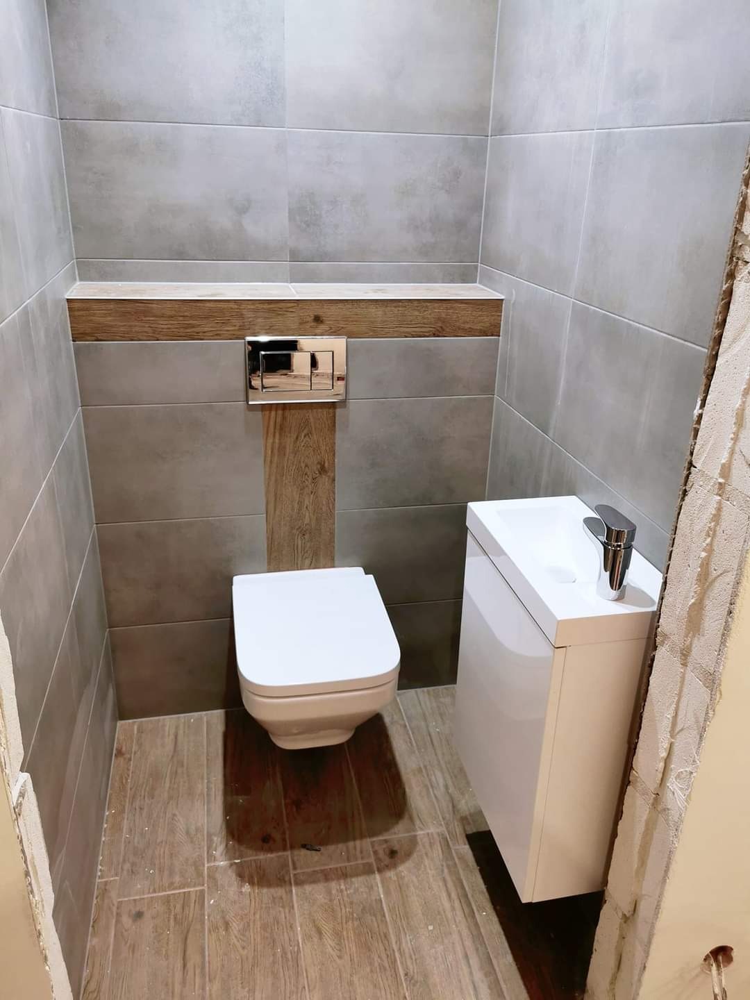 Nowoczesna łazienka w trakcie remontu: szare płytki ścienne i podłogowe imitujące drewno, podwieszana toaleta, biała szafka z umywalką i chromowaną baterią, fragment surowej ściany z widocznym...