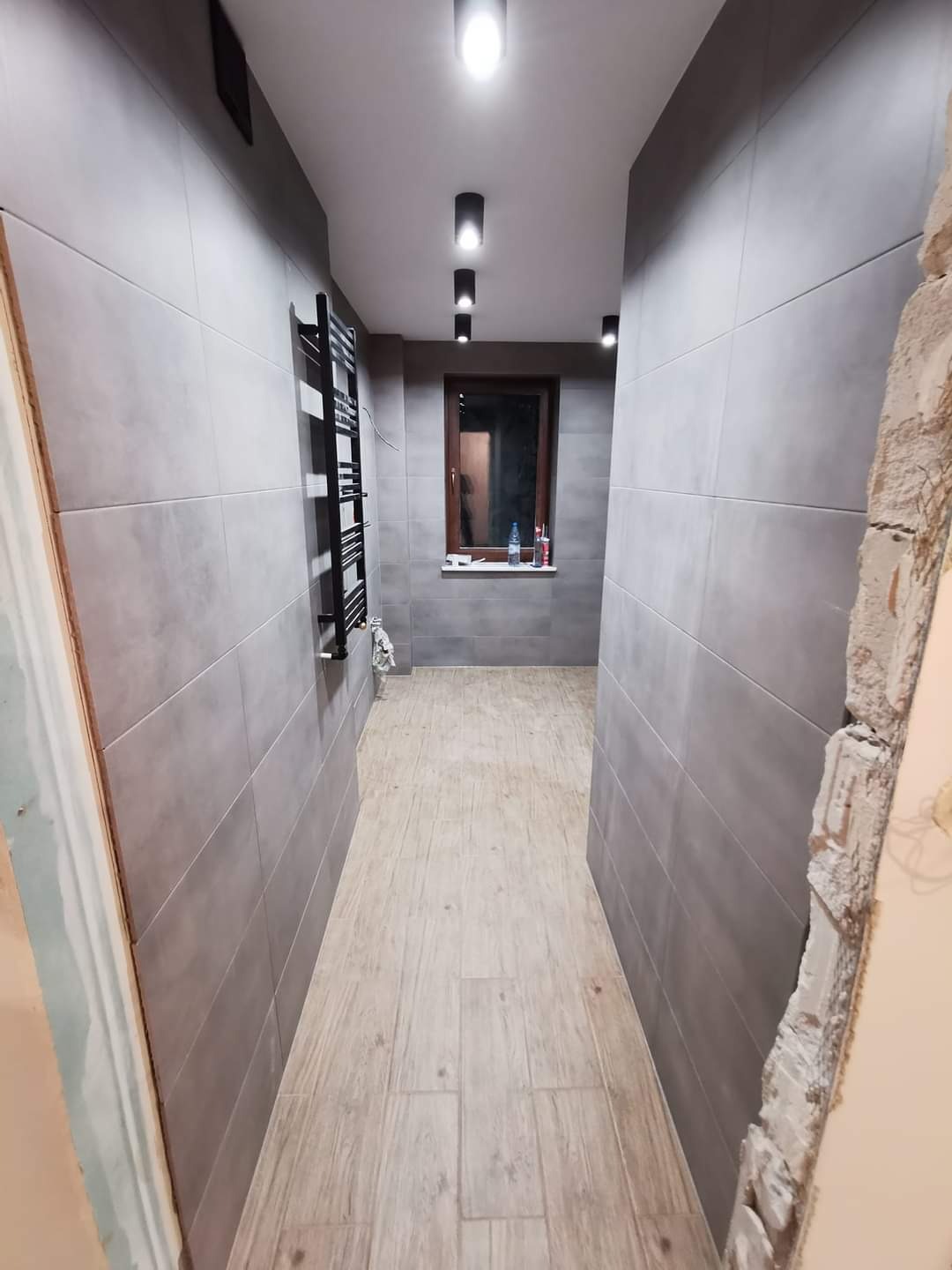 Wąski korytarz łazienkowy z szarymi płytkami ściennymi, drewnopodobną podłogą, czarnym grzejnikiem drabinkowym i oknem z brązową ramą. Widoczny fragment niezakończonego wykończenia ściany przy...