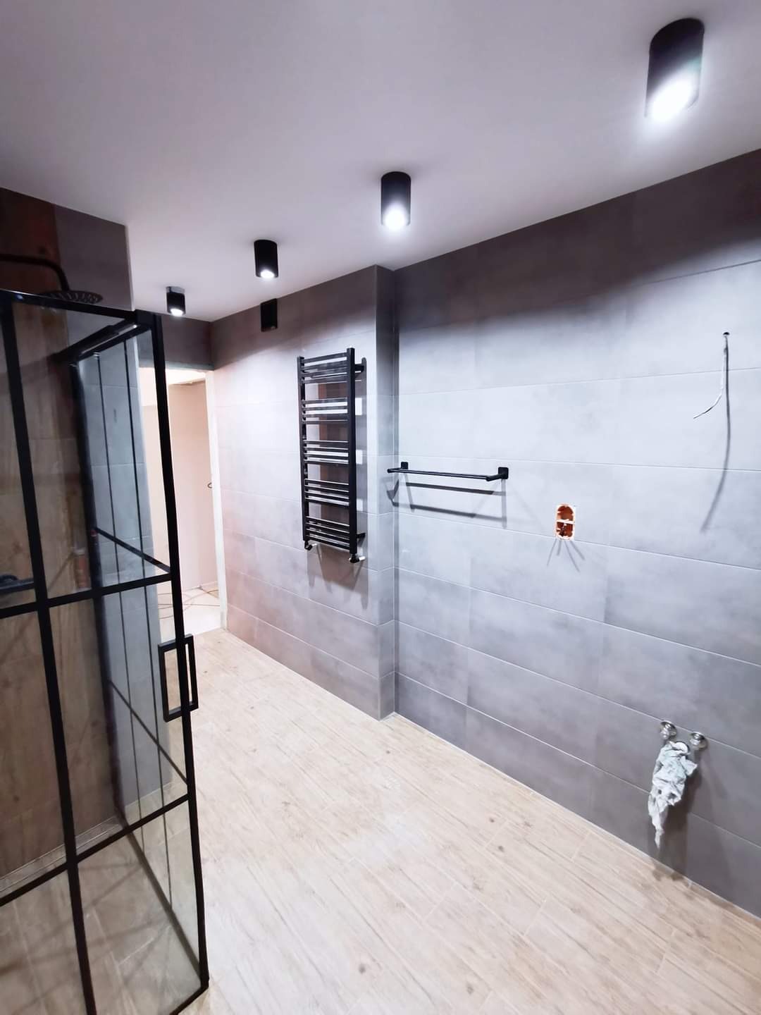 Nowoczesna łazienka z szarymi płytkami, drewnopodobną podłogą, czarną kabiną prysznicową typu walk-in i grzejnikiem drabinkowym, widoczne przygotowane punkty instalacyjne.