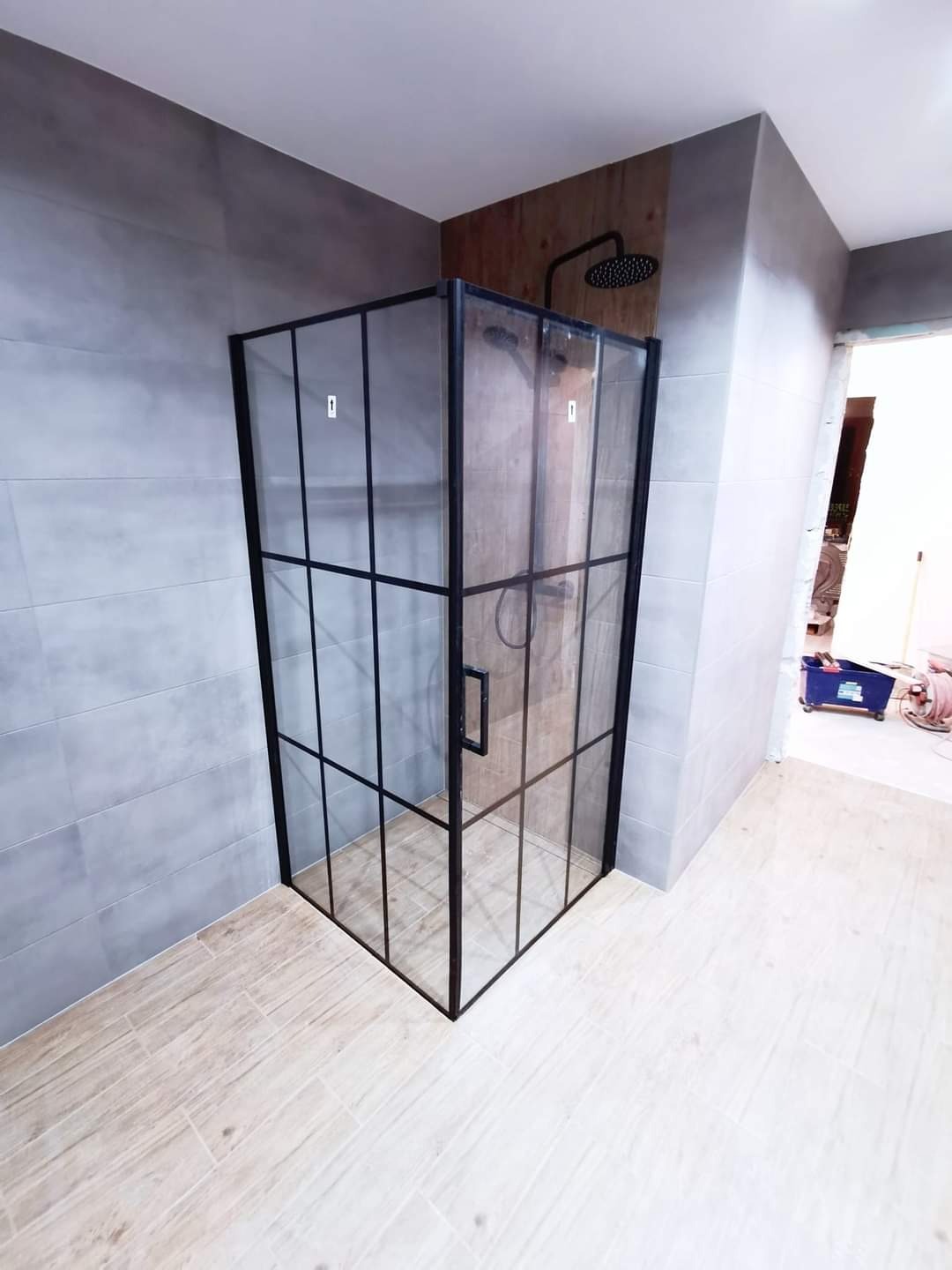 Nowoczesna kabina prysznicowa z czarnymi profilami i transparentnym szkłem, zamontowana w łazience z szarymi płytkami ściennymi i podłogą imitującą drewno.