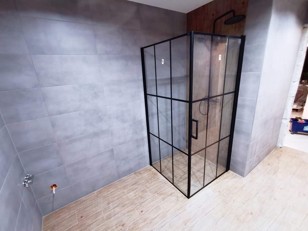 Nowoczesna kabina prysznicowa z czarnymi profilami i szklanymi ściankami, zamontowana w łazience z szarymi płytkami ściennymi i podłogą imitującą drewno. Widoczna deszczownica w kolorze czarnym.