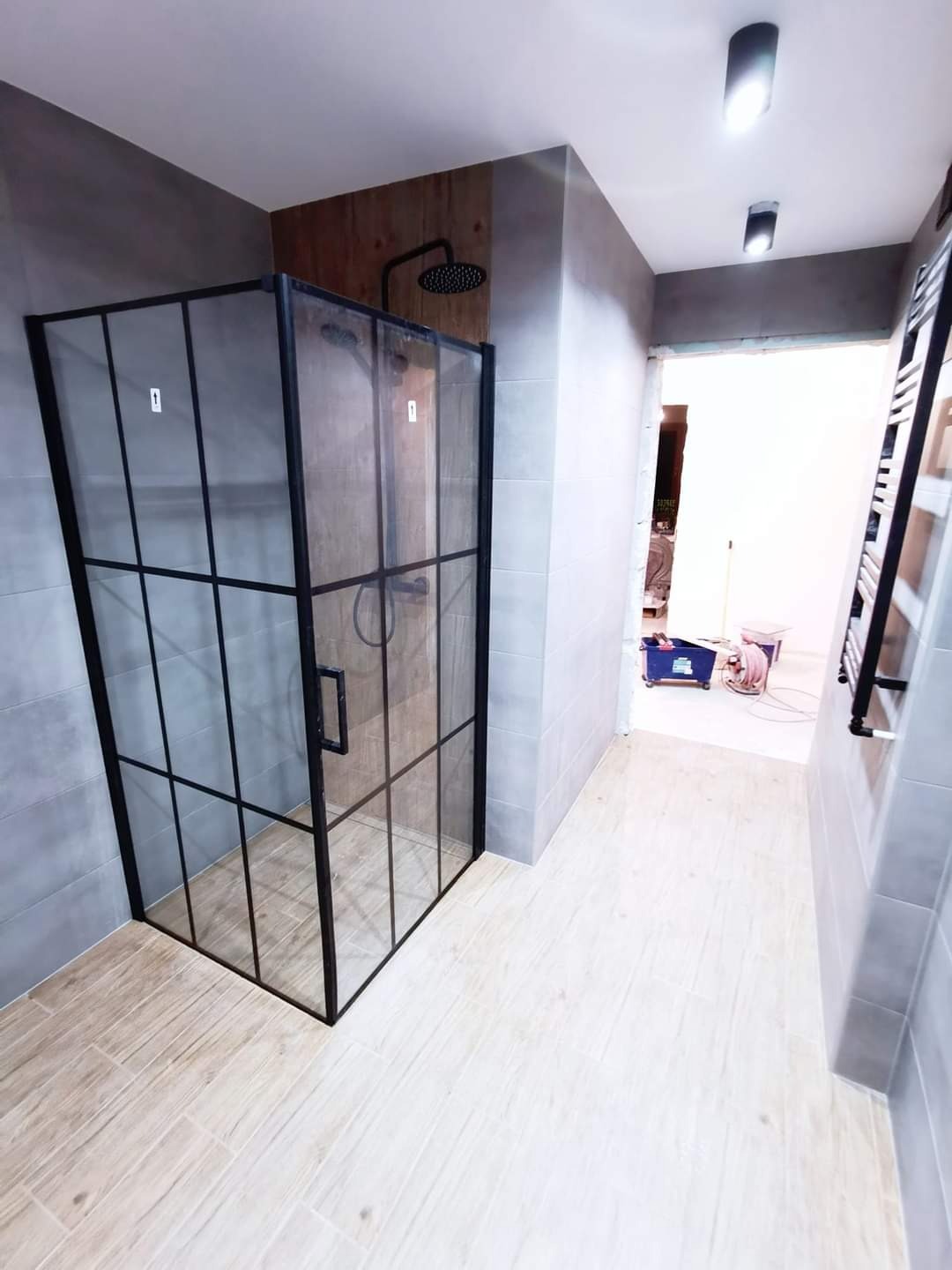 Nowoczesna kabina prysznicowa z czarnymi profilami i szklanymi ściankami, zamontowana w łazience wykończonej szarymi płytkami i drewnopodobną ścianą z deszczownicą.