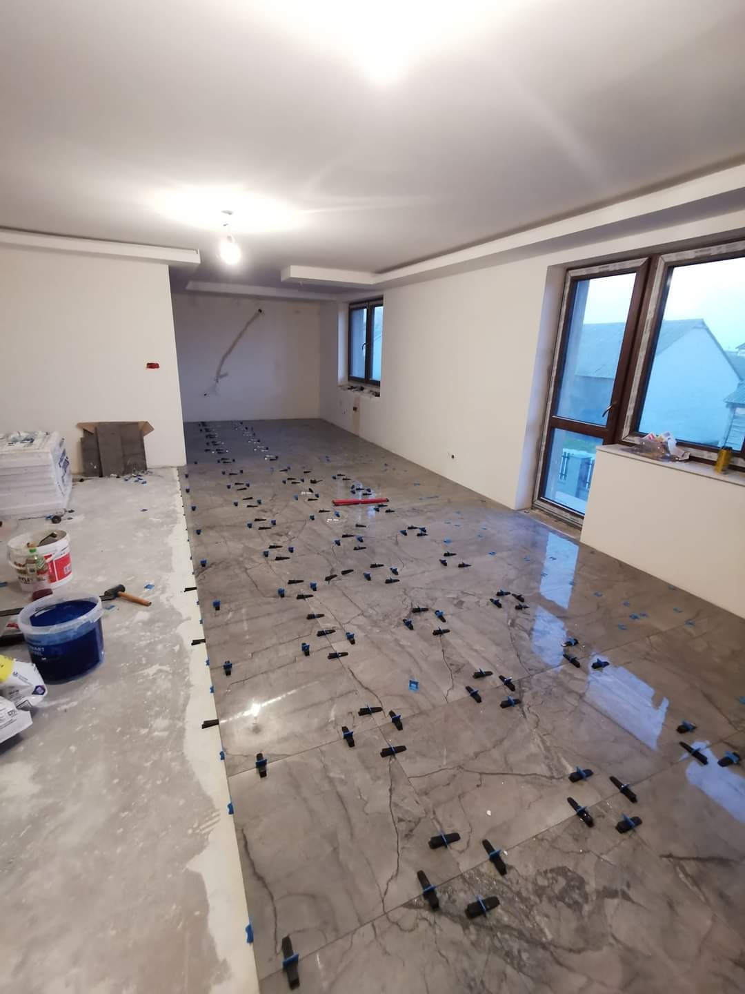 Układanie płytek imitujących marmur na podłodze w dużym pokoju, widoczne niebieskie krzyżyki dystansowe, częściowo położone płytki, wiadro z klejem, paczki z płytkami w tle.