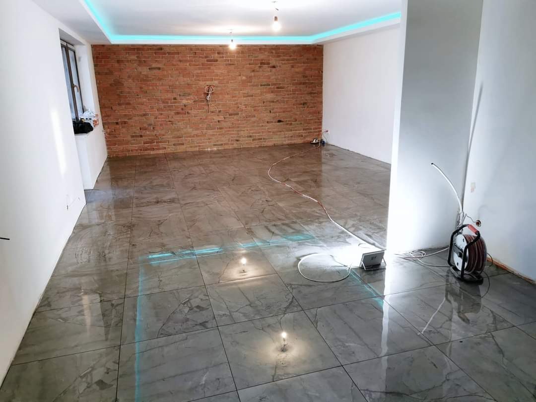 Wnętrze pokoju po remoncie z nową marmurową podłogą, ceglaną ścianą akcentującą i oświetleniem LED pod sufitem. Widoczne narzędzia i lampa budowlana.