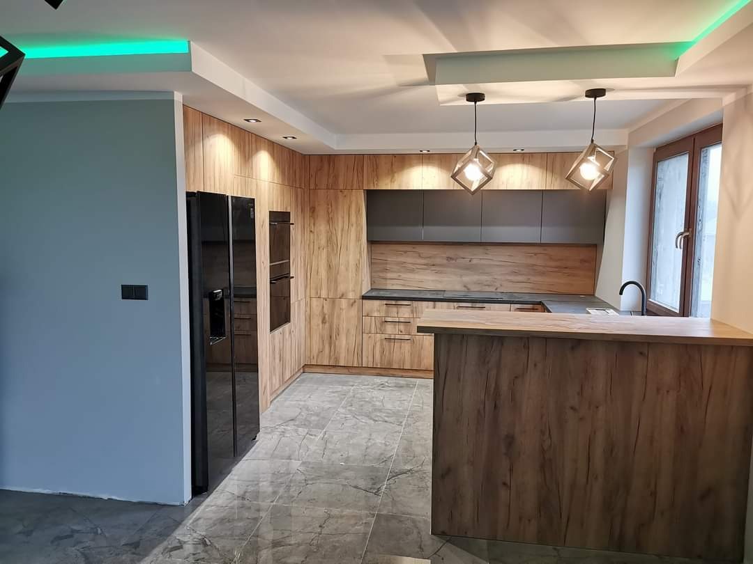 Nowoczesna kuchnia z drewnianymi frontami szafek, czarnym sprzętem AGD i marmurową podłogą, oświetlona geometrycznymi lampami wiszącymi i ledowym światłem sufitowym.