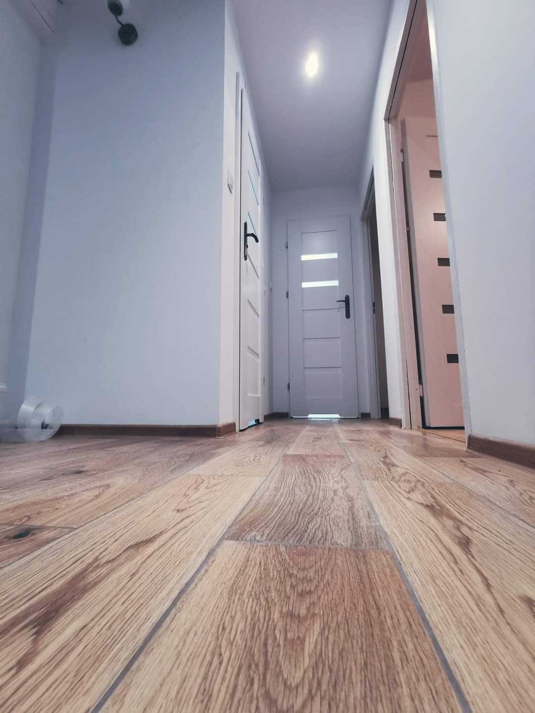 Perspektywa z podłogi na świeżo ułożony parkiet w korytarzu z białymi drzwiami i listwami przypodłogowymi, widoczne ślady po folii ochronnej.