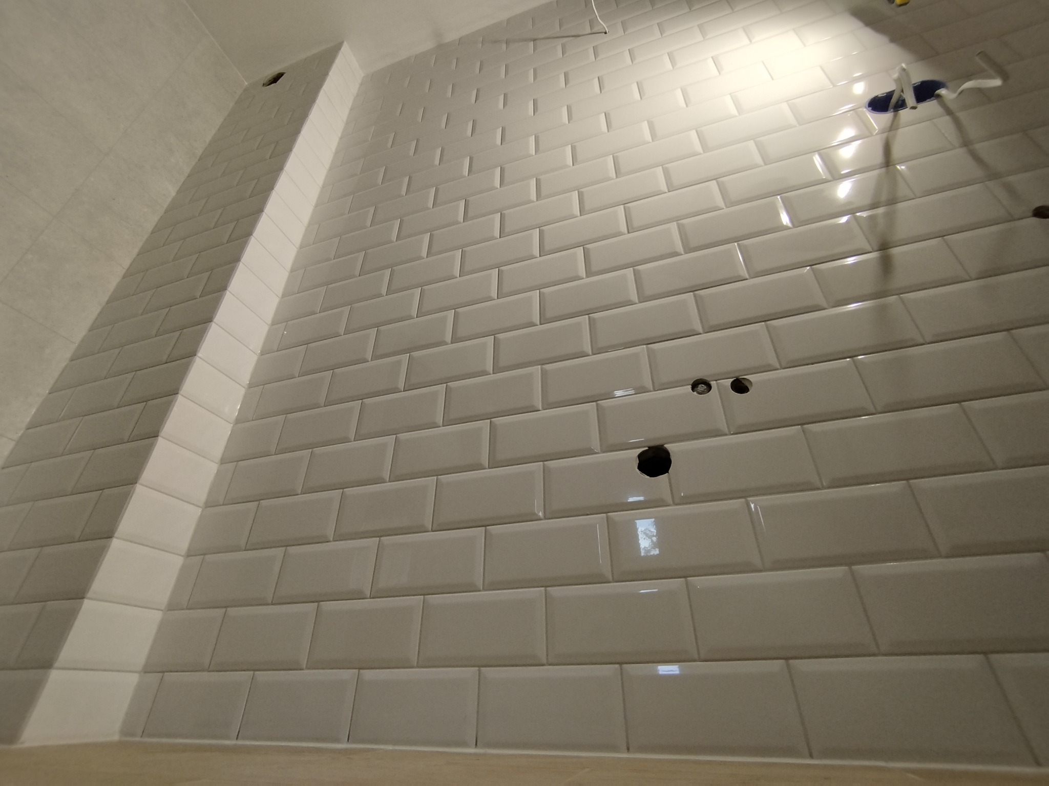 Ściana wyłożona białymi, błyszczącymi kafelkami typu metro, z widocznymi otworami na gniazdka elektryczne i rurę, w trakcie montażu instalacji.