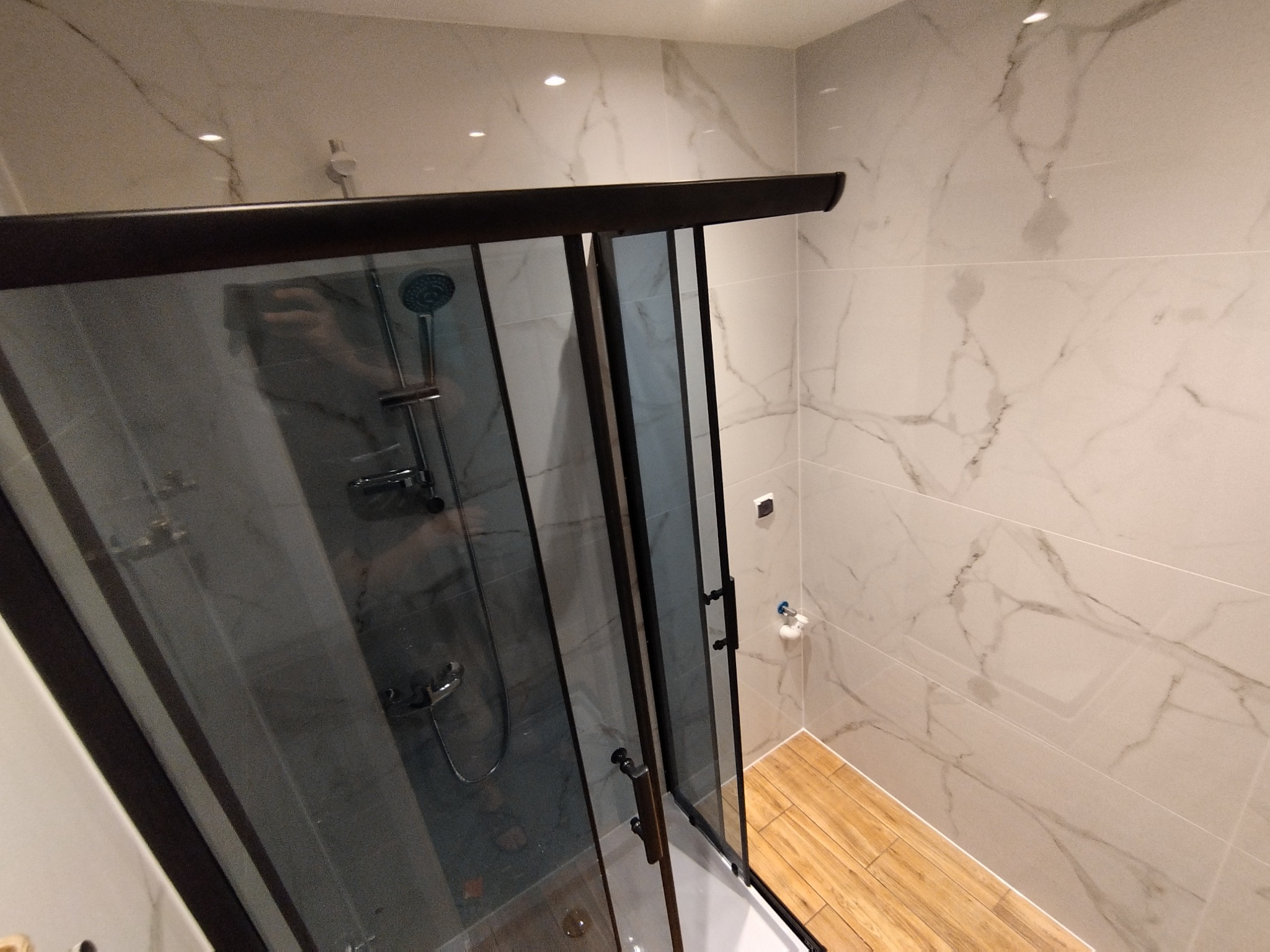 Nowoczesna kabina prysznicowa z ciemną ramą i przesuwnymi drzwiami, widok z góry na marmurowe płytki ścienne i drewnopodobną podłogę.