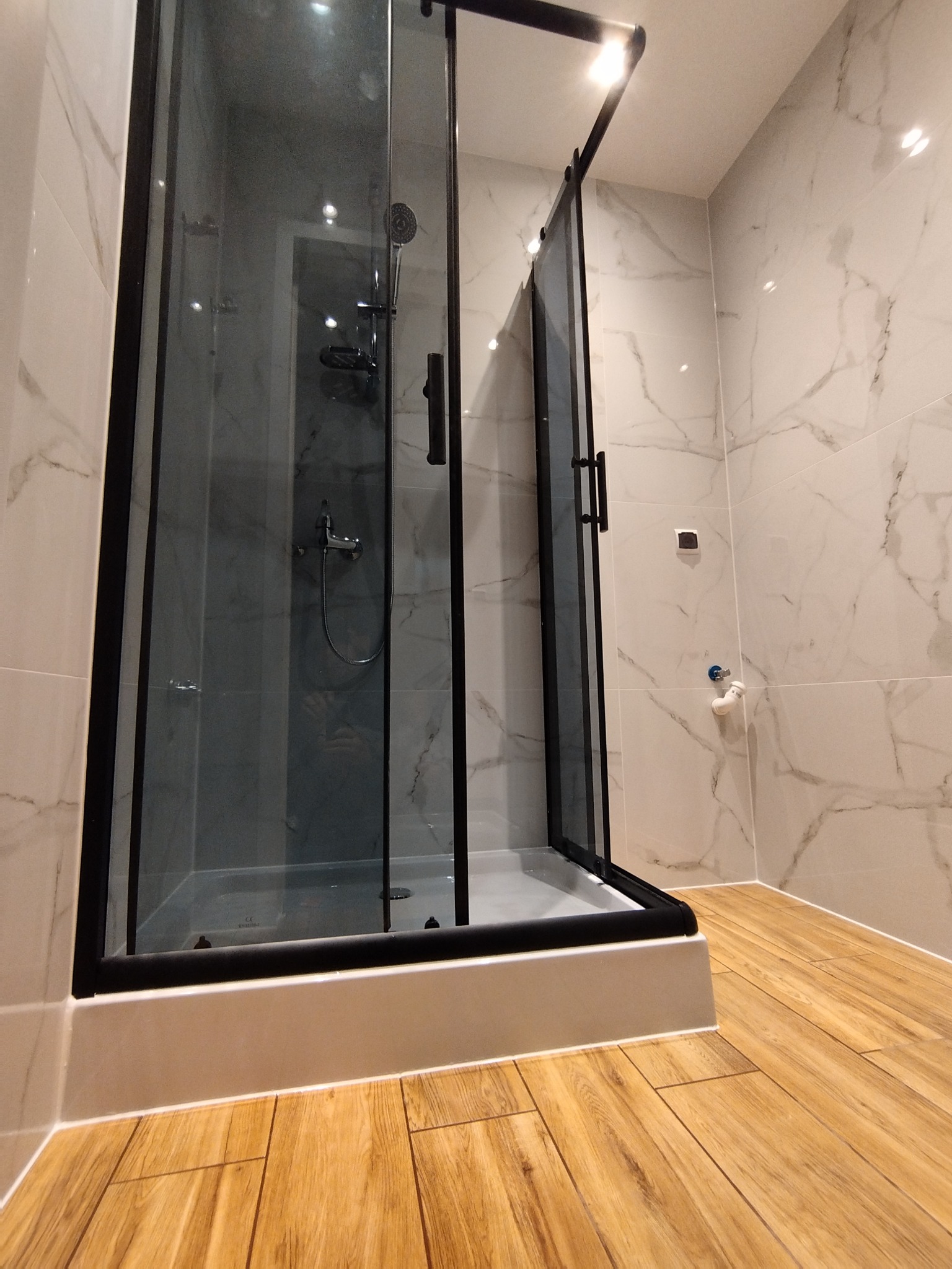 Nowoczesna kabina prysznicowa z czarną ramą i przesuwnymi drzwiami, marmurowe płytki na ścianach i drewnopodobna podłoga w jasnej łazience.
