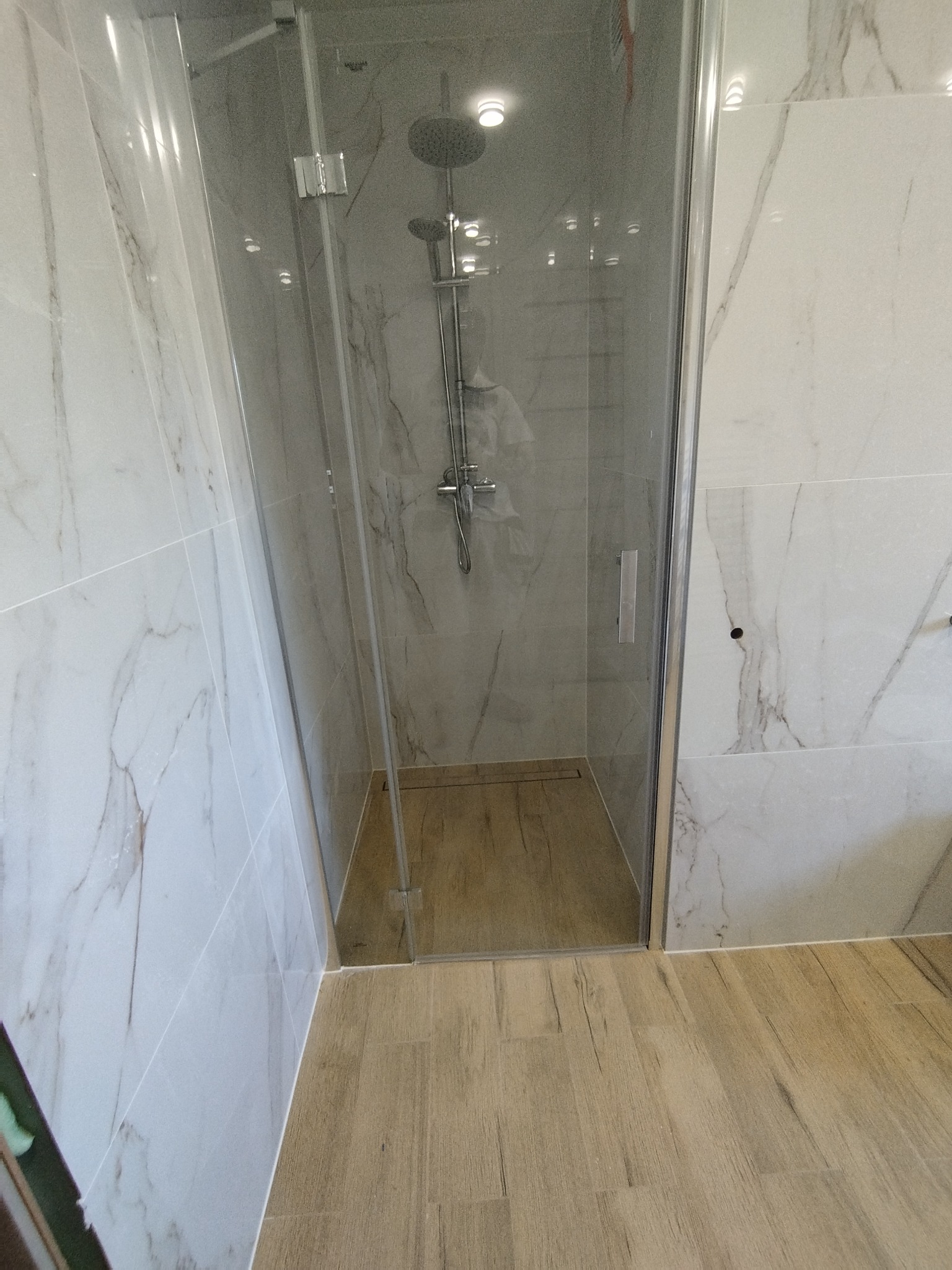 Nowoczesna kabina prysznicowa z deszczownicą, wykończona białymi płytkami imitującymi marmur i podłogą z paneli imitujących drewno.