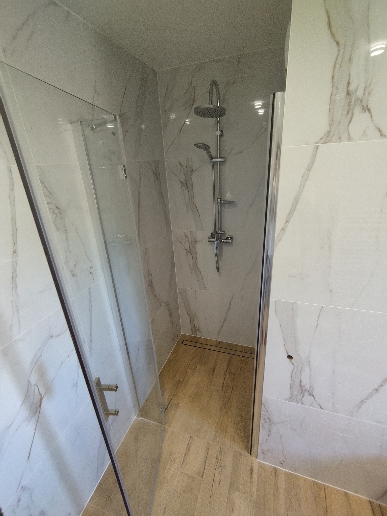 Nowoczesna kabina prysznicowa z deszczownicą i panelami imitującymi marmur, drewnopodobna podłoga, widoczne metalowe uchwyty na szklanej ściance.