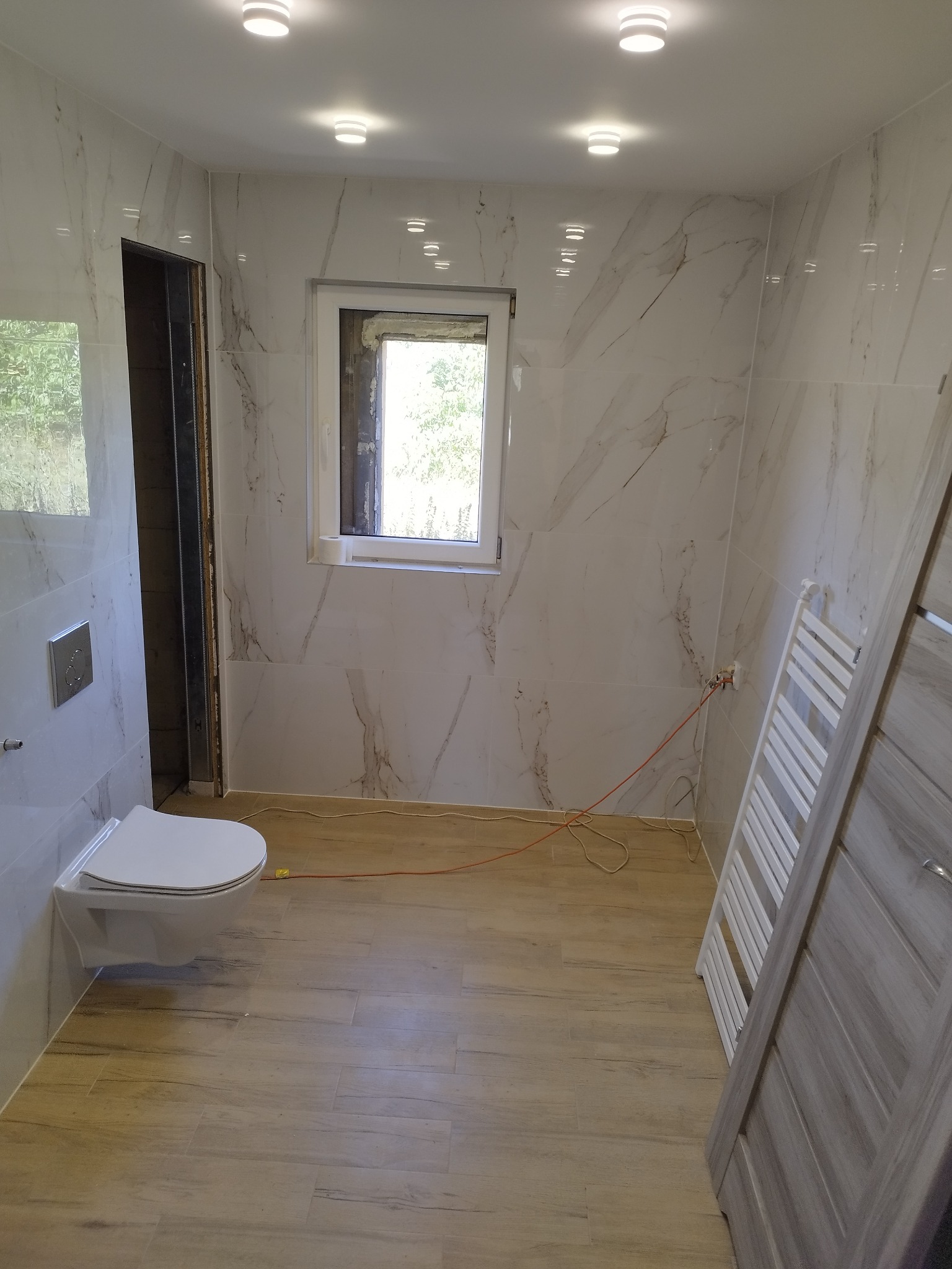 Wnętrze łazienki w trakcie remontu: białe marmurowe płytki ścienne, podwieszana toaleta, grzejnik drabinkowy, drewnopodobne płytki podłogowe, okno z widokiem na zieleń, widoczne przewody elektryczne.