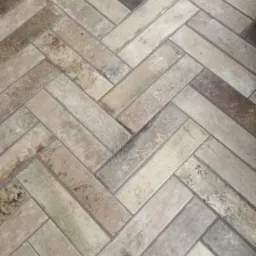 Ułożona w jodełkę podłoga z płytek imitujących cegłę w odcieniach beżu, brązu i szarości, z widoczną fakturą i fugami. Czarny dywanik w rogu kadru.