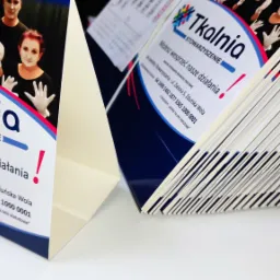 Stos ulotek reklamowych Stowarzyszenia Tkalnia z grafiką przedstawiającą osoby w kostiumach mimów, ułożonych w perspektywie z widocznymi krawędziami papieru.