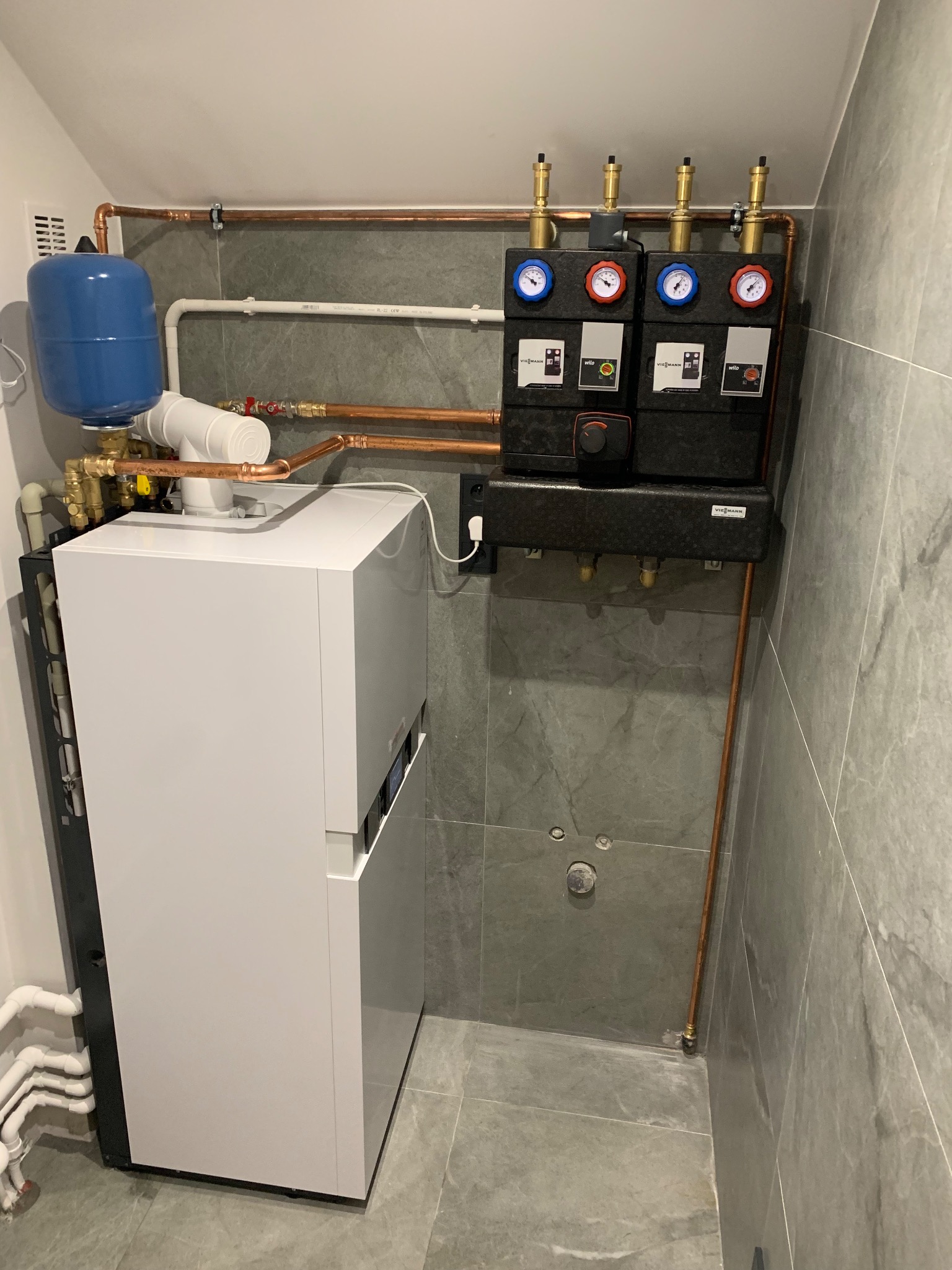 Nowoczesny, biały piec gazowy Viessmann z niebieskim zbiornikiem wyrównawczym i czarnym modułem sterującym z manometrami, zamontowany na tle szarej, marmurowej ściany i podłogi z widocznymi...