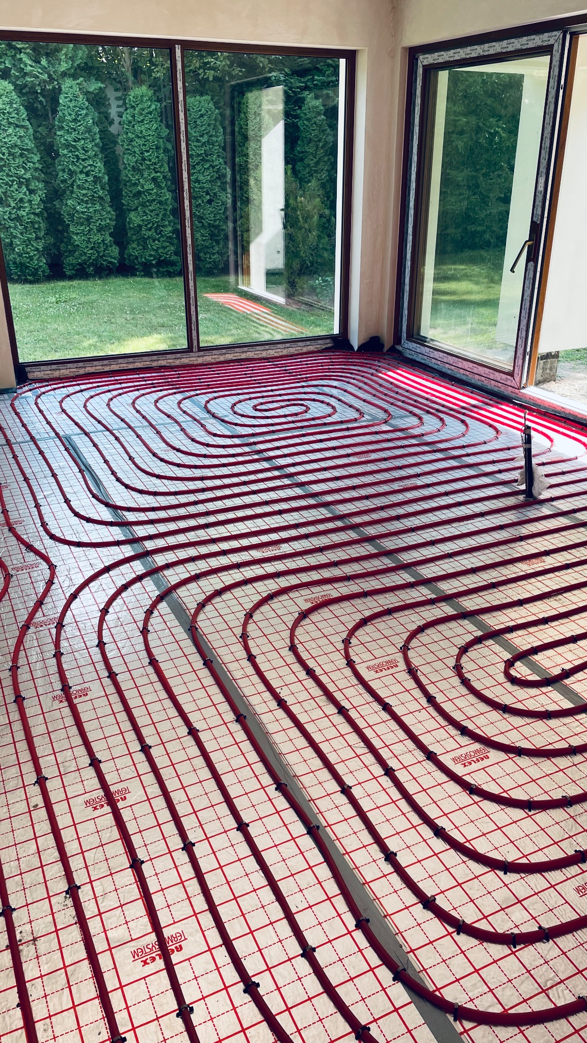 Instalacja ogrzewania podłogowego z czerwonych rur na białej macie z czerwoną siatką w jasnym pomieszczeniu z dużymi oknami wychodzącymi na zielony ogród.