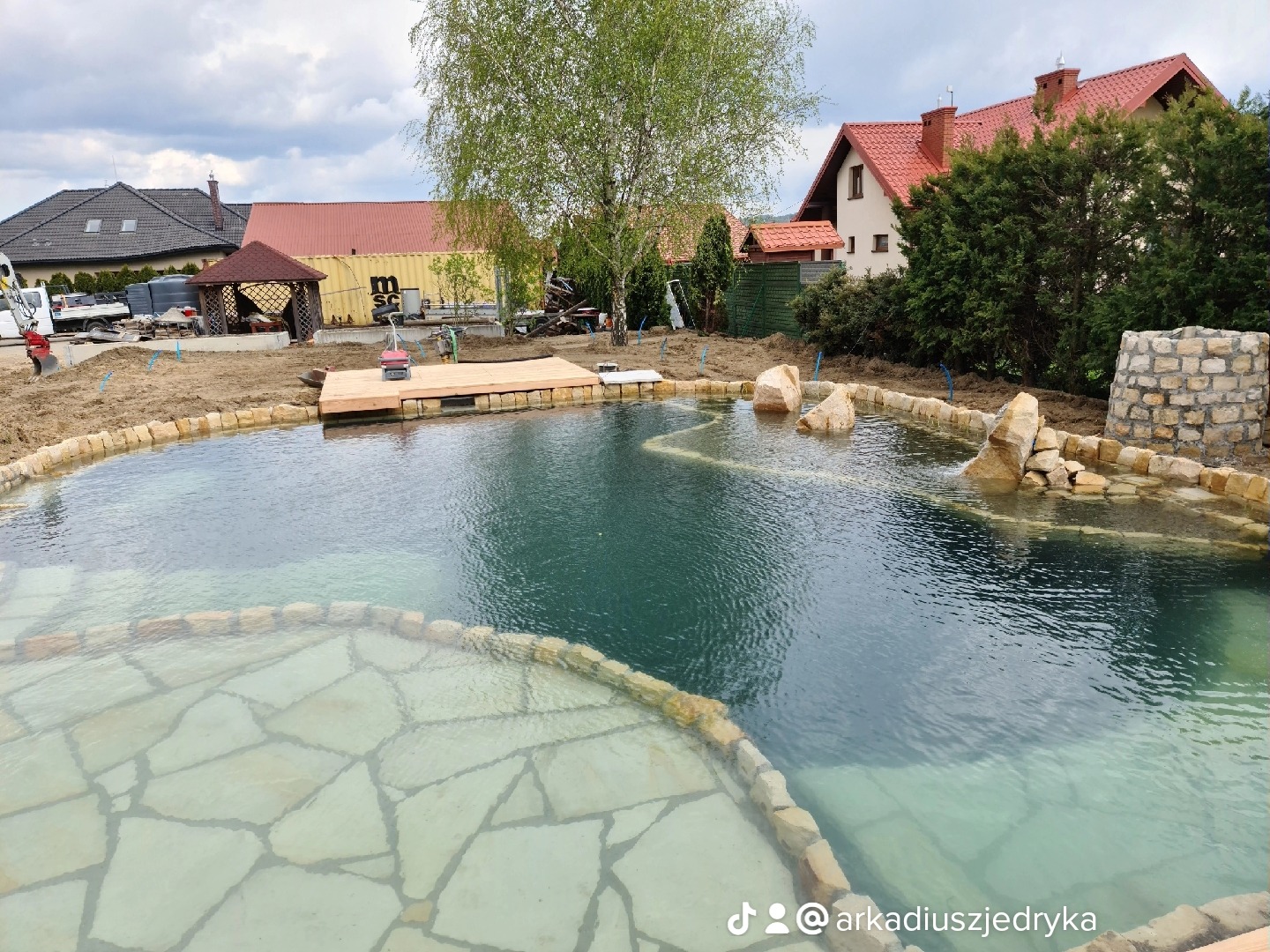 Wykonywanie naturalnego basenu kąpielowego z kamiennym wykończeniem w Tarnowcu, z widoczną strefą płytką i pomostem drewnianym, w tle domy i drzewa.
