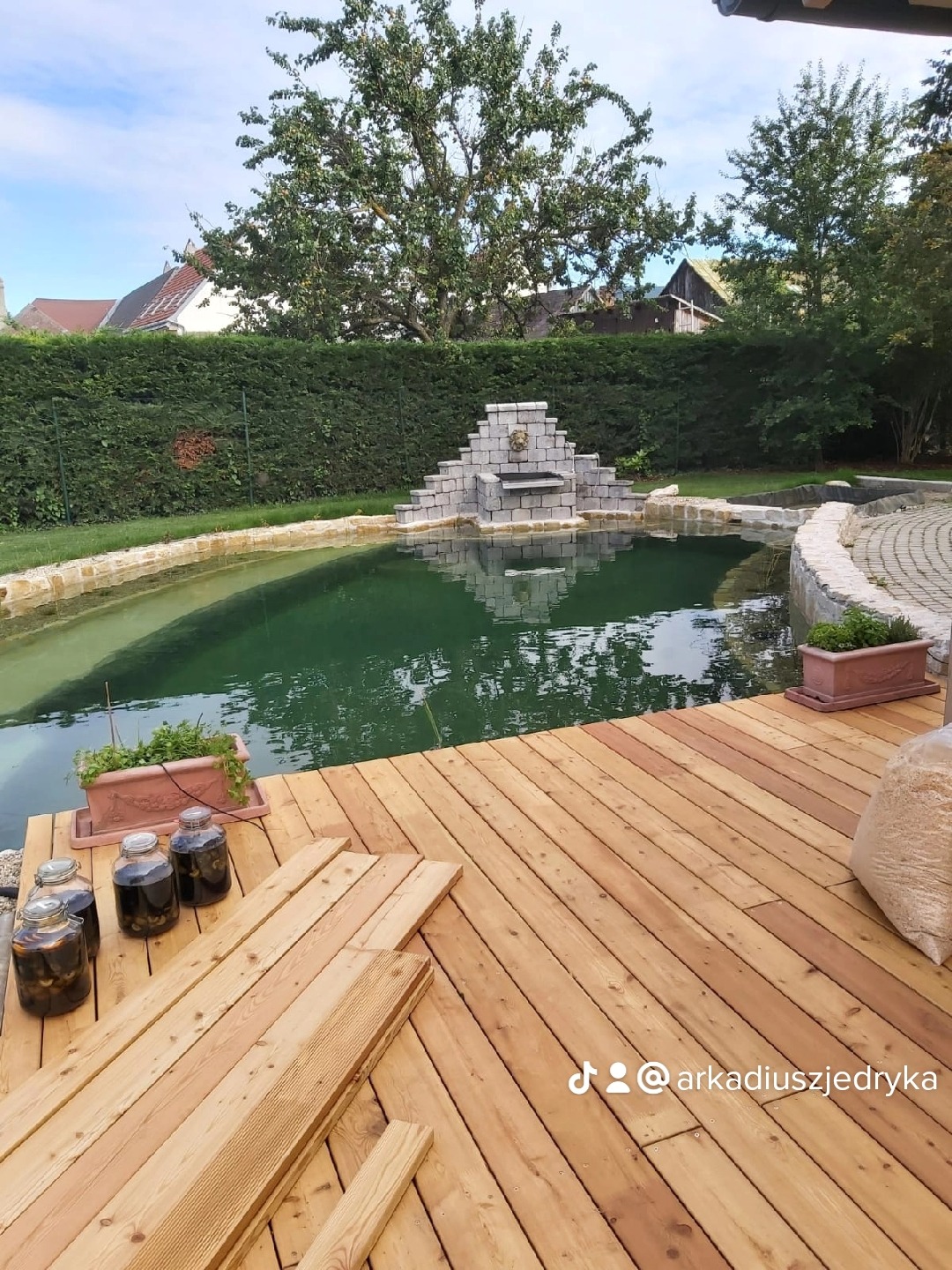 Budowa drewnianego tarasu przy zielonym stawie z kamienną kaskadą i rzeźbą lwa, w tle żywopłot i zabudowania.