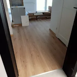 Ułożone panele podłogowe w odcieniach drewna, widoczne z perspektywy progu drzwi, z elementami remontu w tle: zapakowane meble i drzwi.