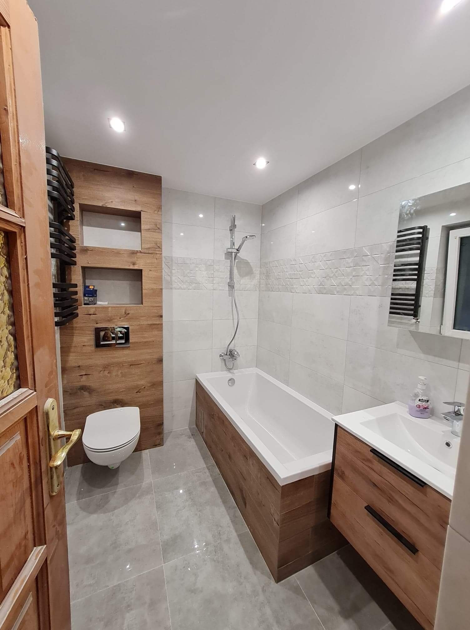 Nowoczesna łazienka z drewnianymi akcentami: obudowa wanny i szafka pod umywalką z drewnopodobnych płytek, płytki imitujące drewno na ścianie z wnękami, szare płytki podłogowe, biała armatura...