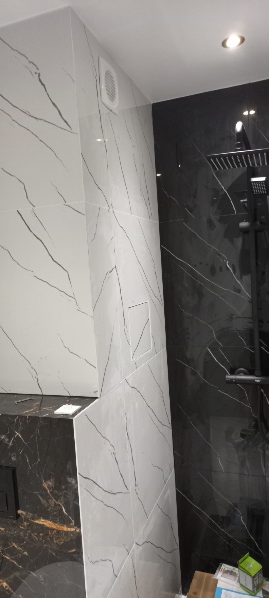 Nowoczesna łazienka z czarno-białymi płytkami imitującymi marmur, widoczny czarny panel prysznicowy i kratka wentylacyjna w górnym rogu.