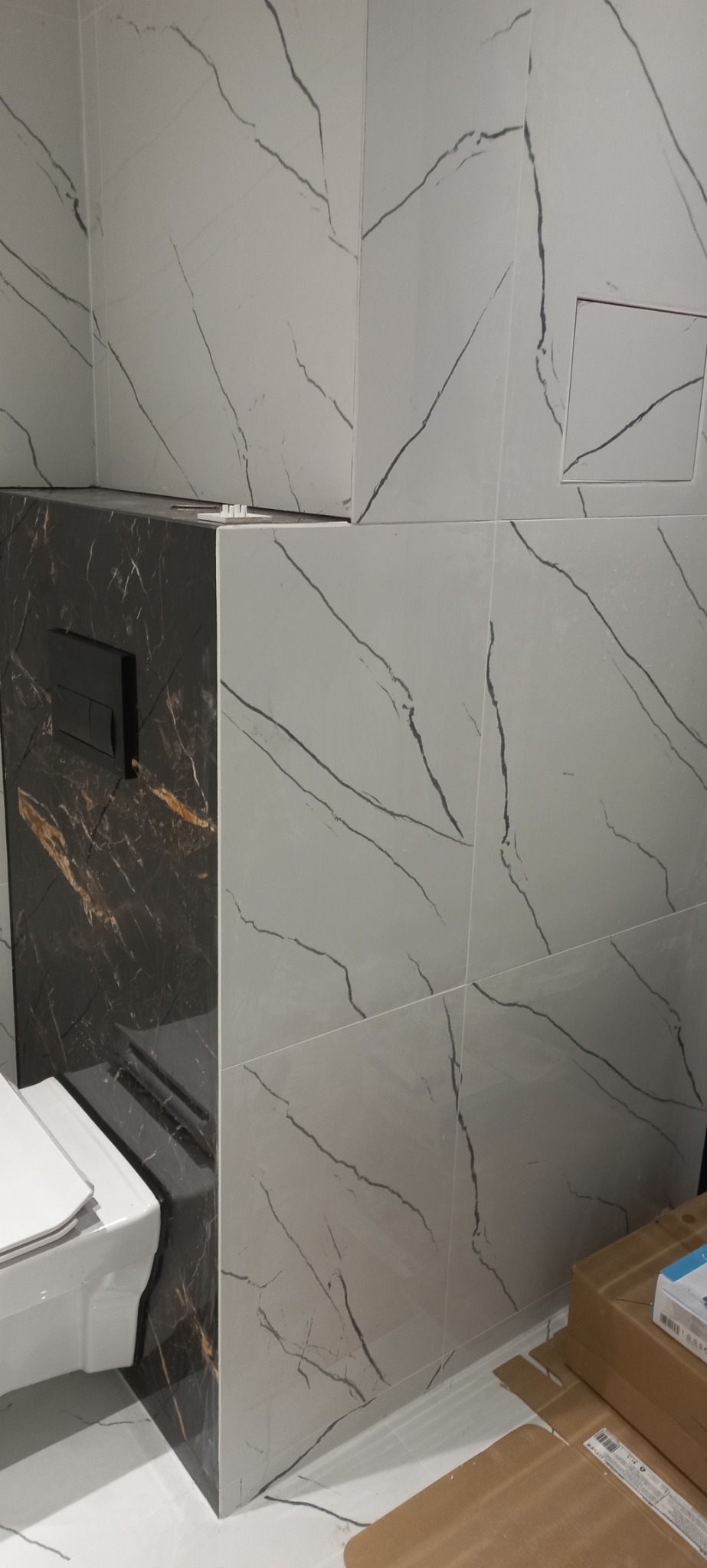 Wykończona łazienka w Łowiczu: białe płytki imitujące marmur na ścianach, zabudowa WC w czarnym marmurze z czarnym przyciskiem spłukującym, widoczny fragment toalety.