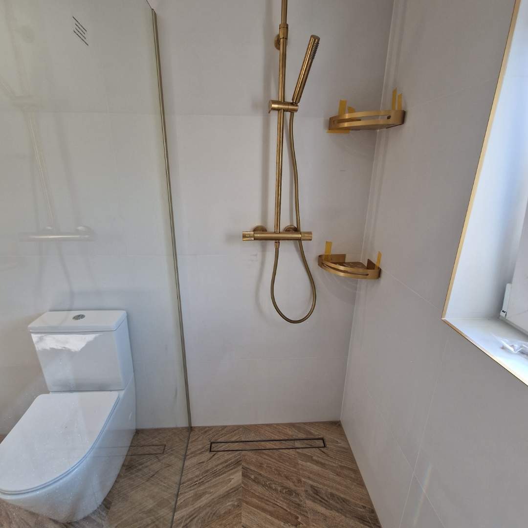 Wykończona łazienka z kabiną prysznicową, toaletą i złotą armaturą. Na ścianach białe płytki, na podłodze wzór jodełki. Widoczny odpływ liniowy.