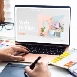 Projektant stron internetowych używa tabletu graficznego do tworzenia wizualizacji na laptopie z wyświetloną stroną web design, obok paleta kolorów i okulary.