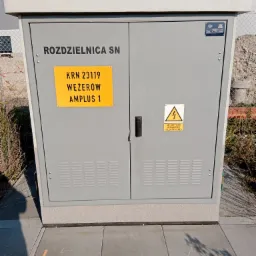 Szara, zewnętrzna rozdzielnica SN z żółtą tabliczką adresową KRN 23119 Węzerów Amplus 1 i znakiem ostrzegawczym przed porażeniem elektrycznym, umieszczona na betonowej podstawie obok chodnika...
