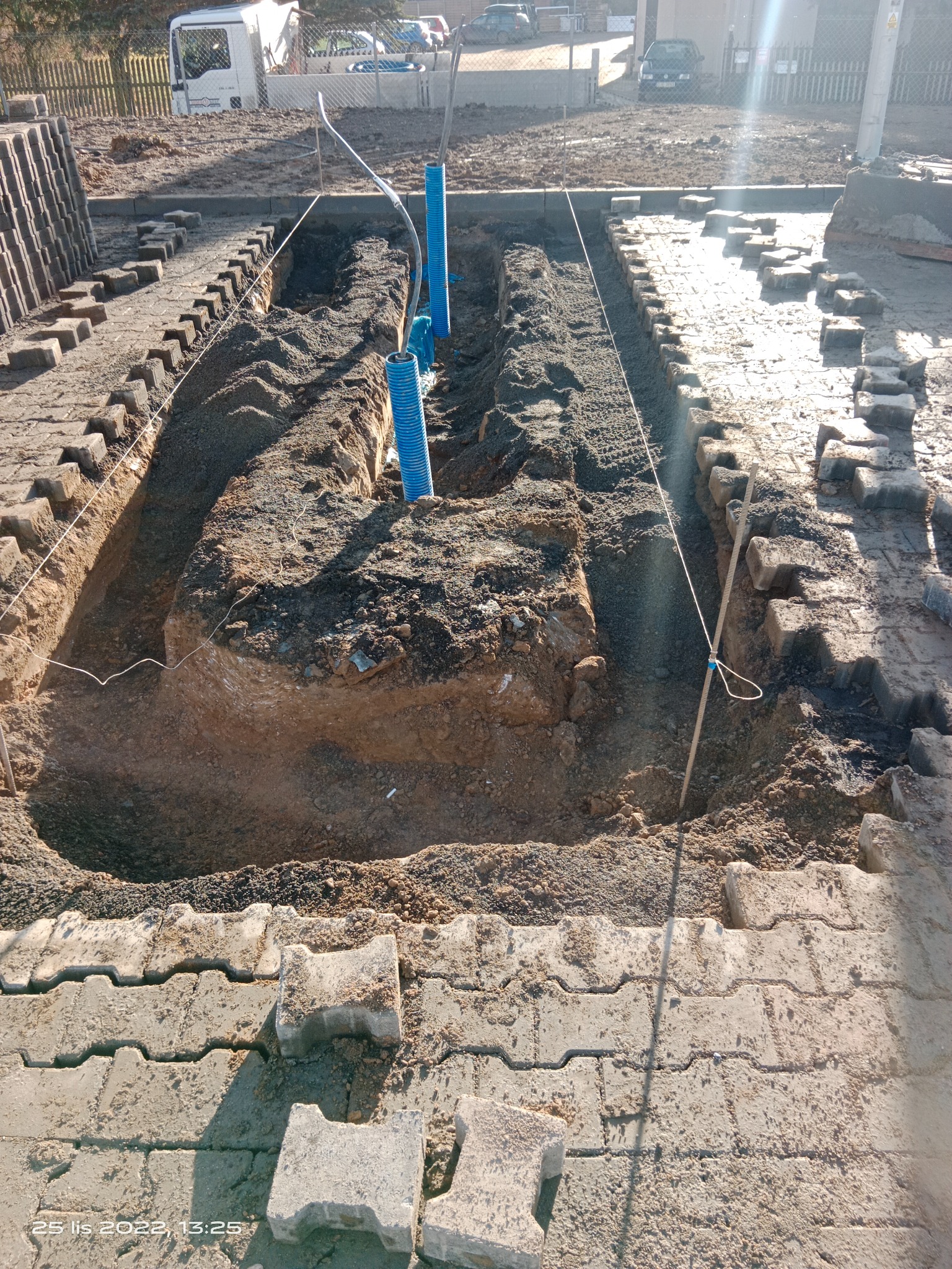 Wykop pod instalację z widocznymi niebieskimi rurami osłonowymi, obrzeża z kostki brukowej, częściowo ułożone, w tle budynek i samochody.