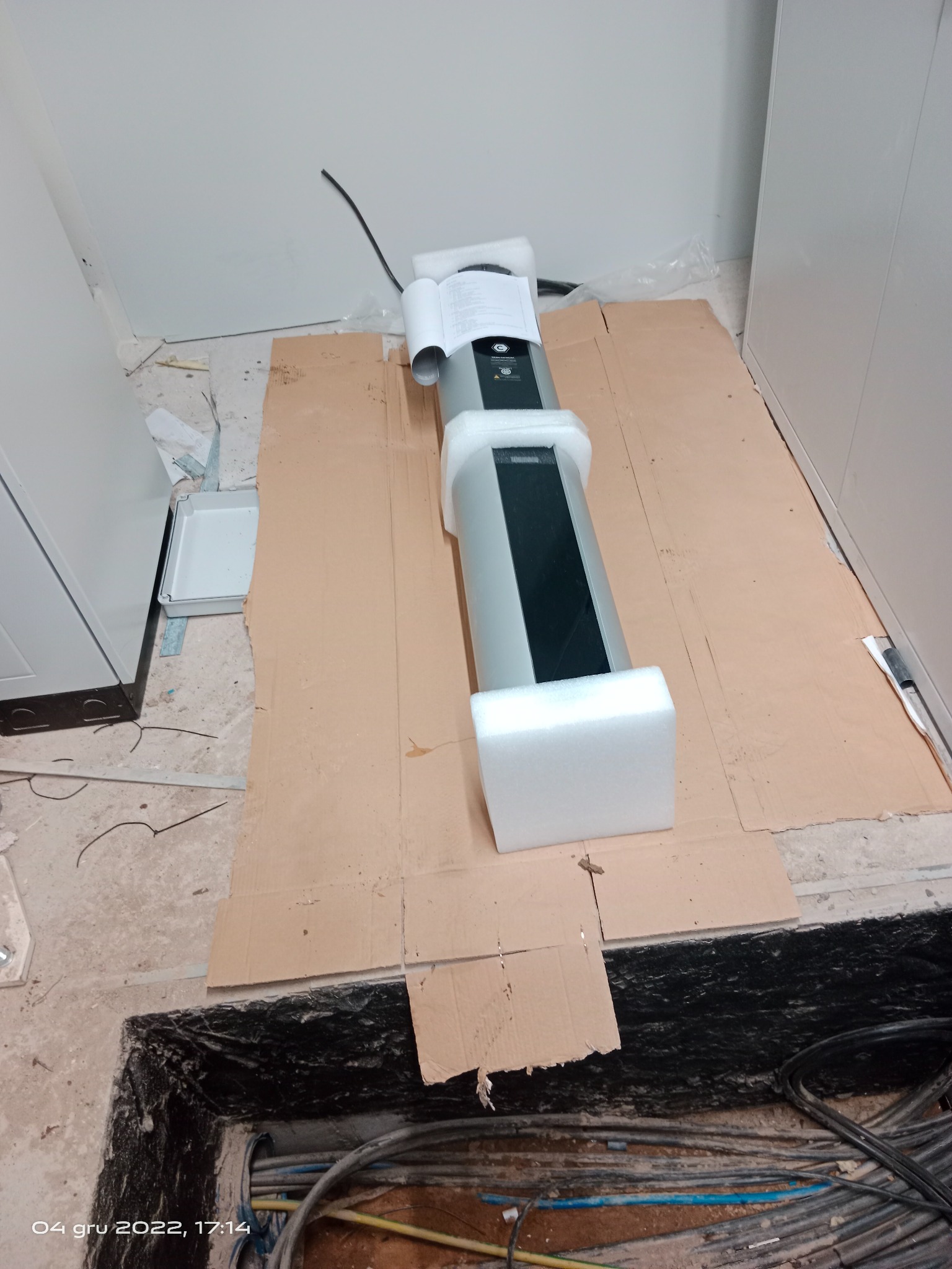 Nowoczesna, srebrna skrzynka elektryczna w ochronnym opakowaniu styropianowym, umieszczona na kartonie na podłodze w trakcie instalacji, z widocznymi kablami w otworze pod podłogą.