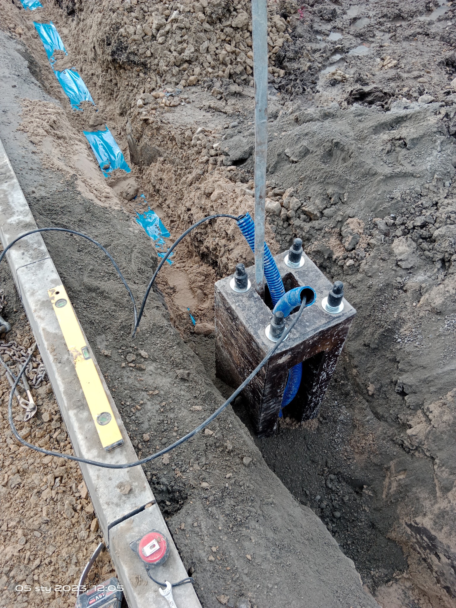 Wykop z ułożonymi rurami osłonowymi w niebieskiej folii, skrzynka z wyprowadzonymi rurami karbowanymi, poziomica i miarka na krawężniku, widok z góry.