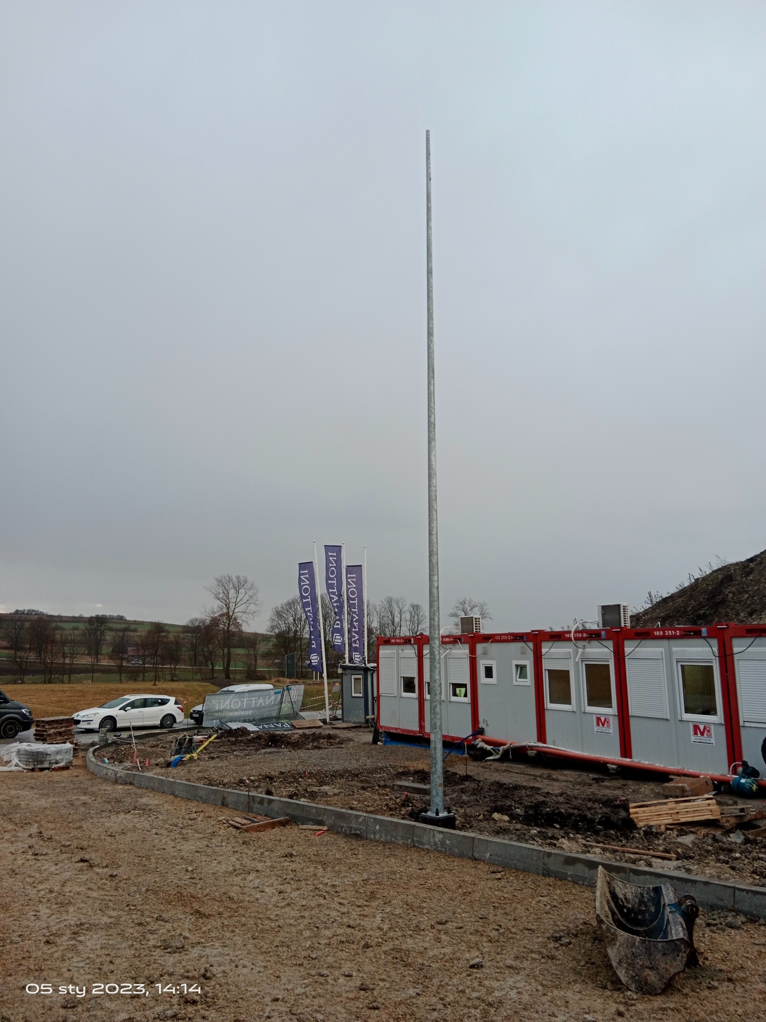 Stalowy maszt na placu budowy, w tle kontenery biurowe i firmowe flagi, pochmurne niebo.