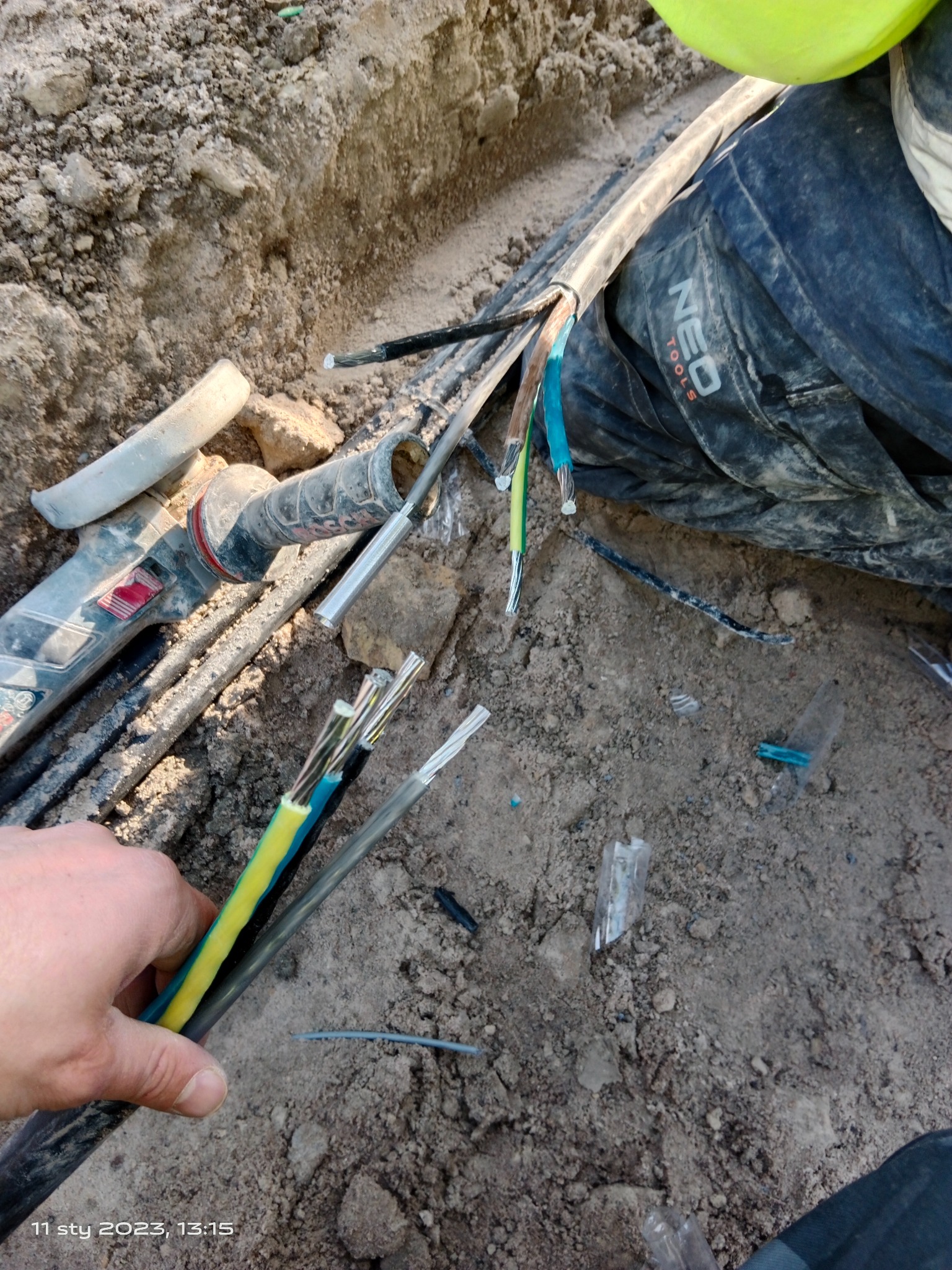 Podłączenie kabli elektrycznych w wykopie, widoczne narzędzia Bosch i fragment roboczego ubrania marki Neo Tools.