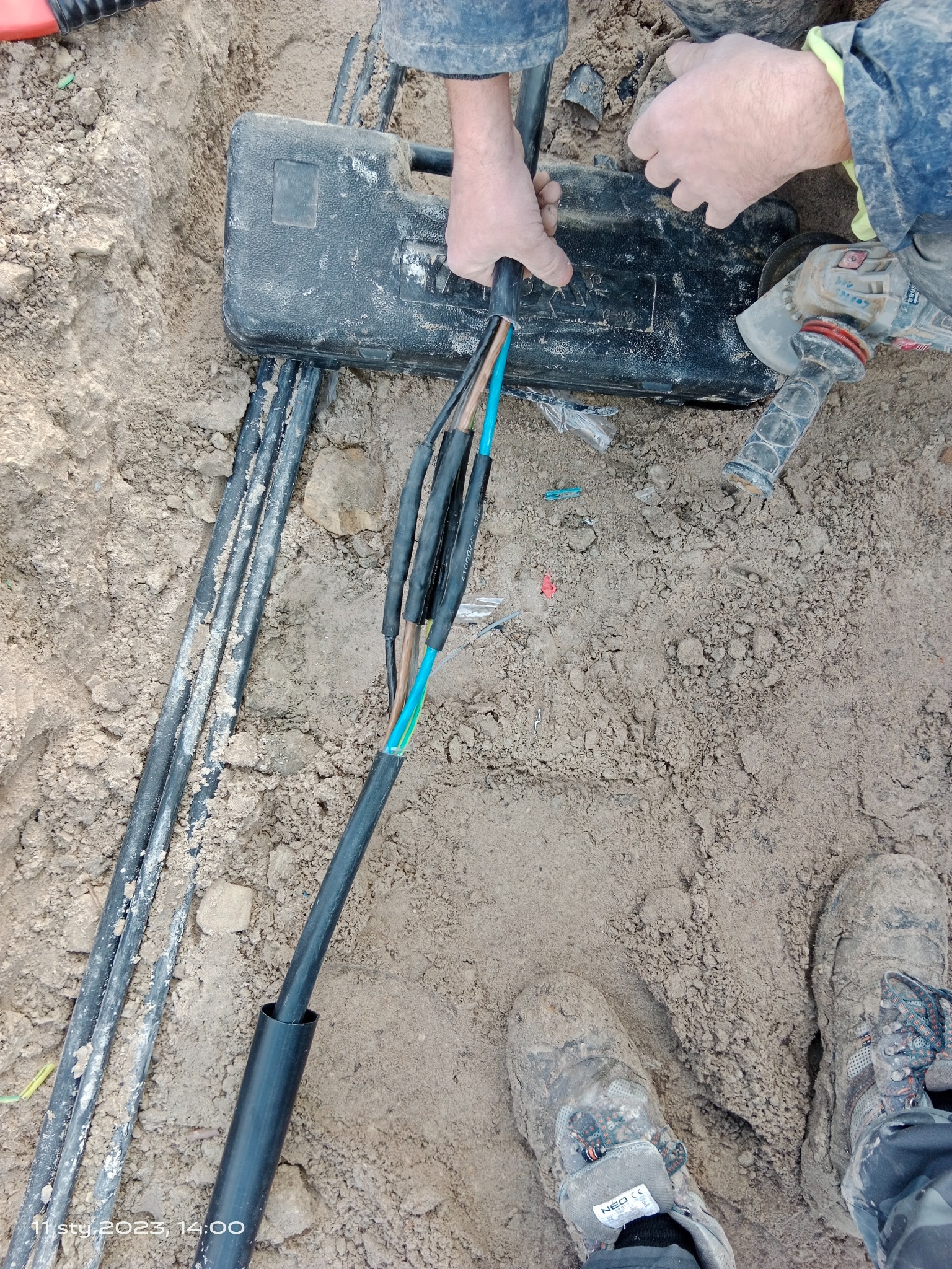 Robocze ujęcie z ziemi: ręce elektryka łączące kable w ziemi obok czarnej skrzynki narzędziowej i szlifierki kątowej, widoczne brudne buty robocze.